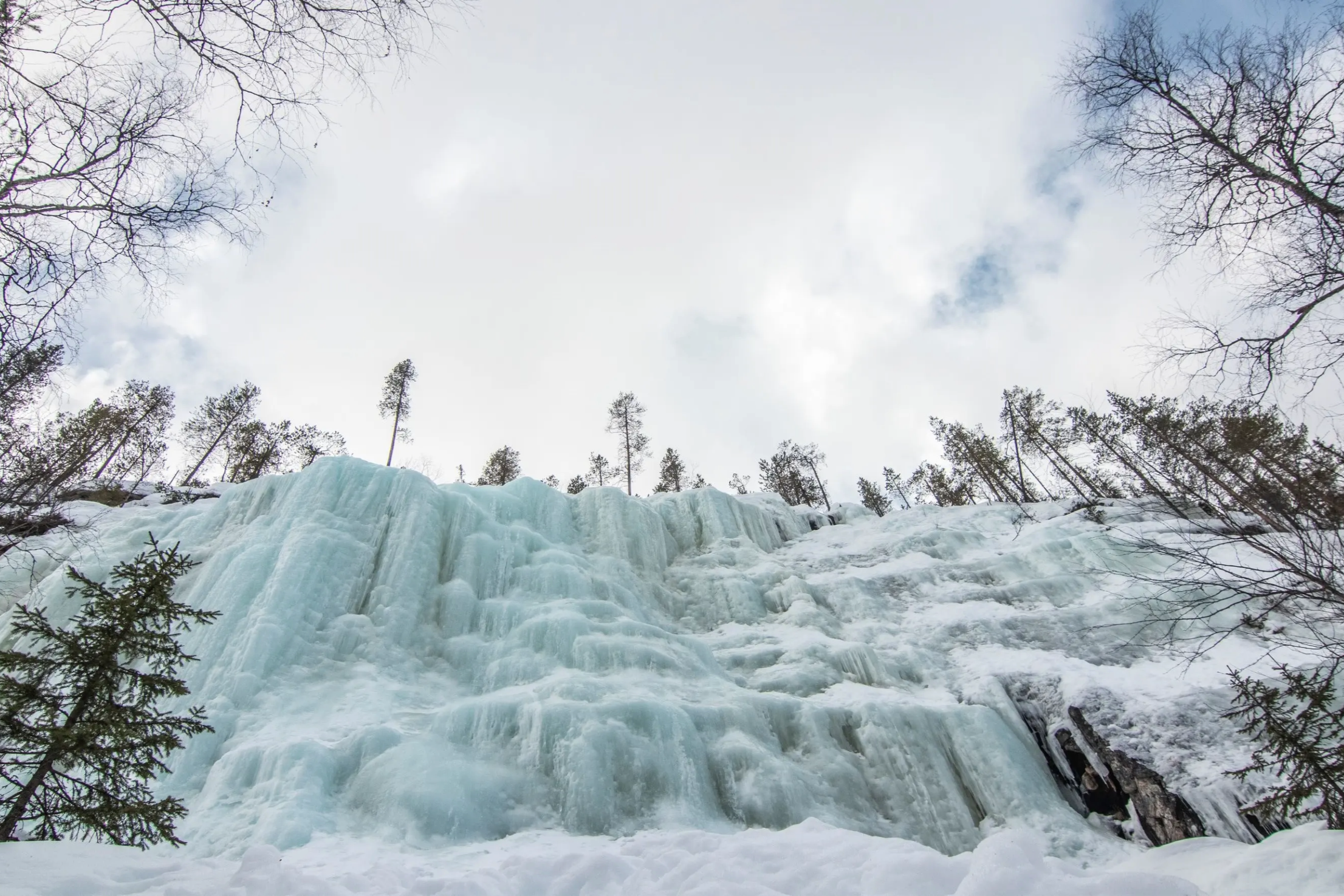 Ice-covered waterfall surrounded by snow and trees, viewed from the ground looking upward toward a cloudy sky.