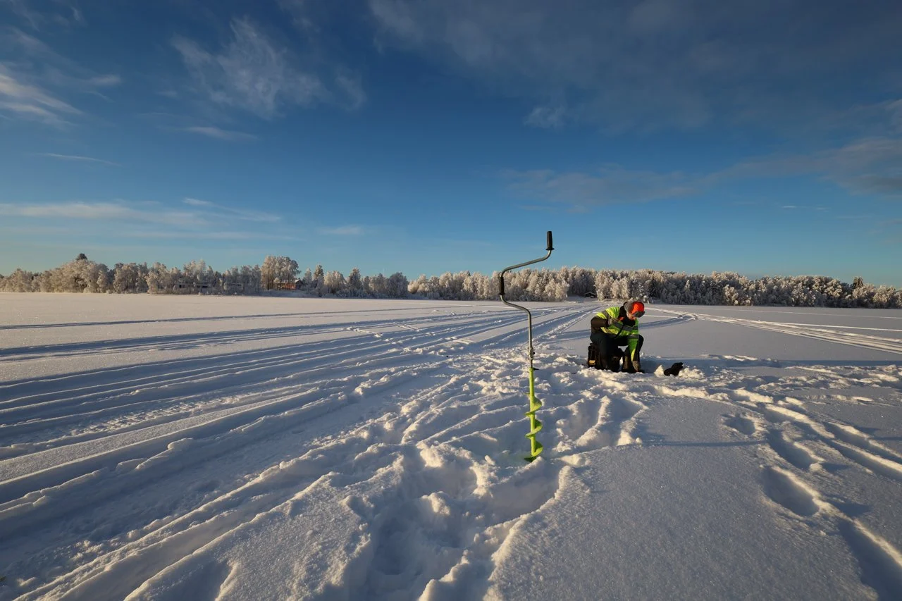 A person in winter gear kneels next to a backpack on a snow-covered field, with a snow auger nearby, under a partly cloudy blue sky.