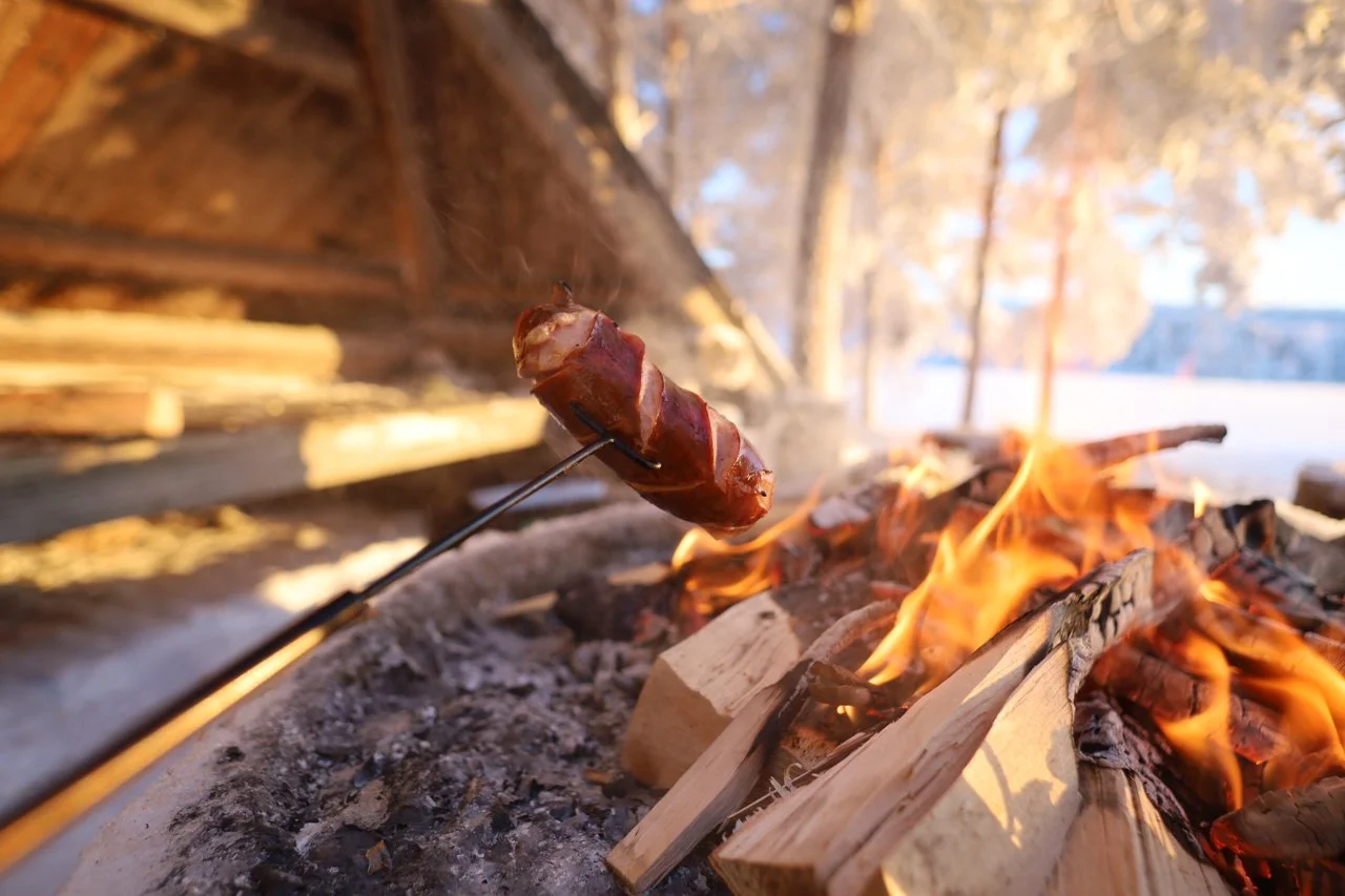 A sausage on a stick over a campfire with burning wood, outdoors.