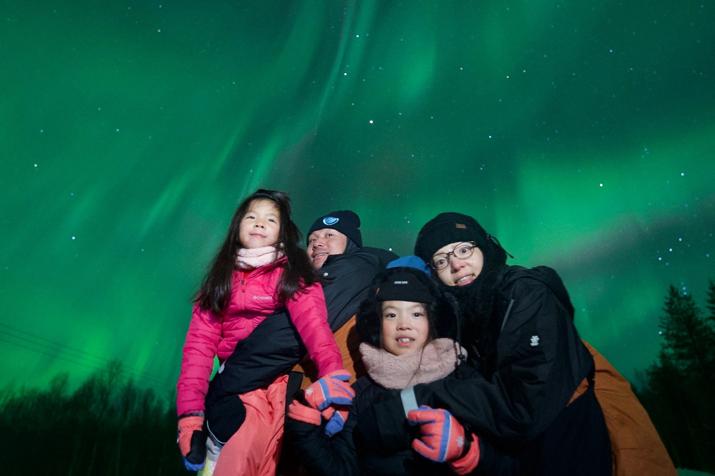 Family of four dressed in winter gear, standing outdoors at night under the green Northern Lights with starry sky in the background.