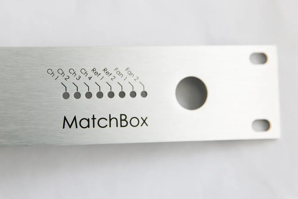 A metal MatchBox audio equipment component with labeled input/output connections for Ch 1 to Ch 4, Refl, Fan 1, Fan 2, and a large hole in the center.