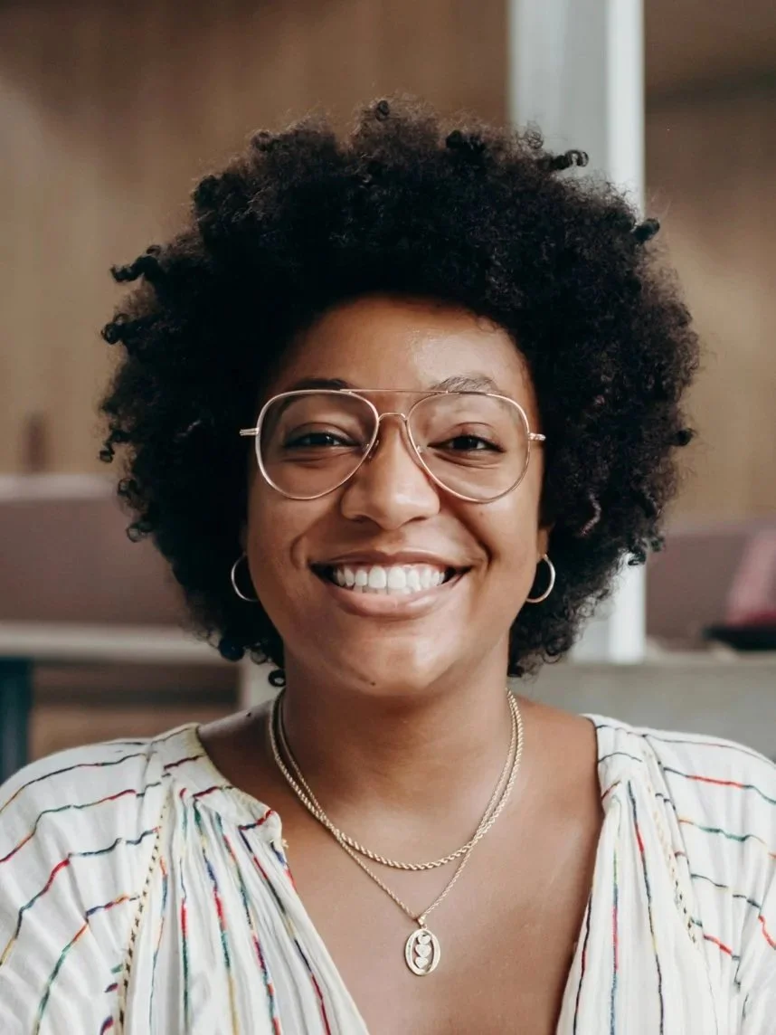 A woman with a big curly afro hairstyle smiling, wearing glasses, hoop earrings, layered necklaces, and a striped blouse in an indoor setting.