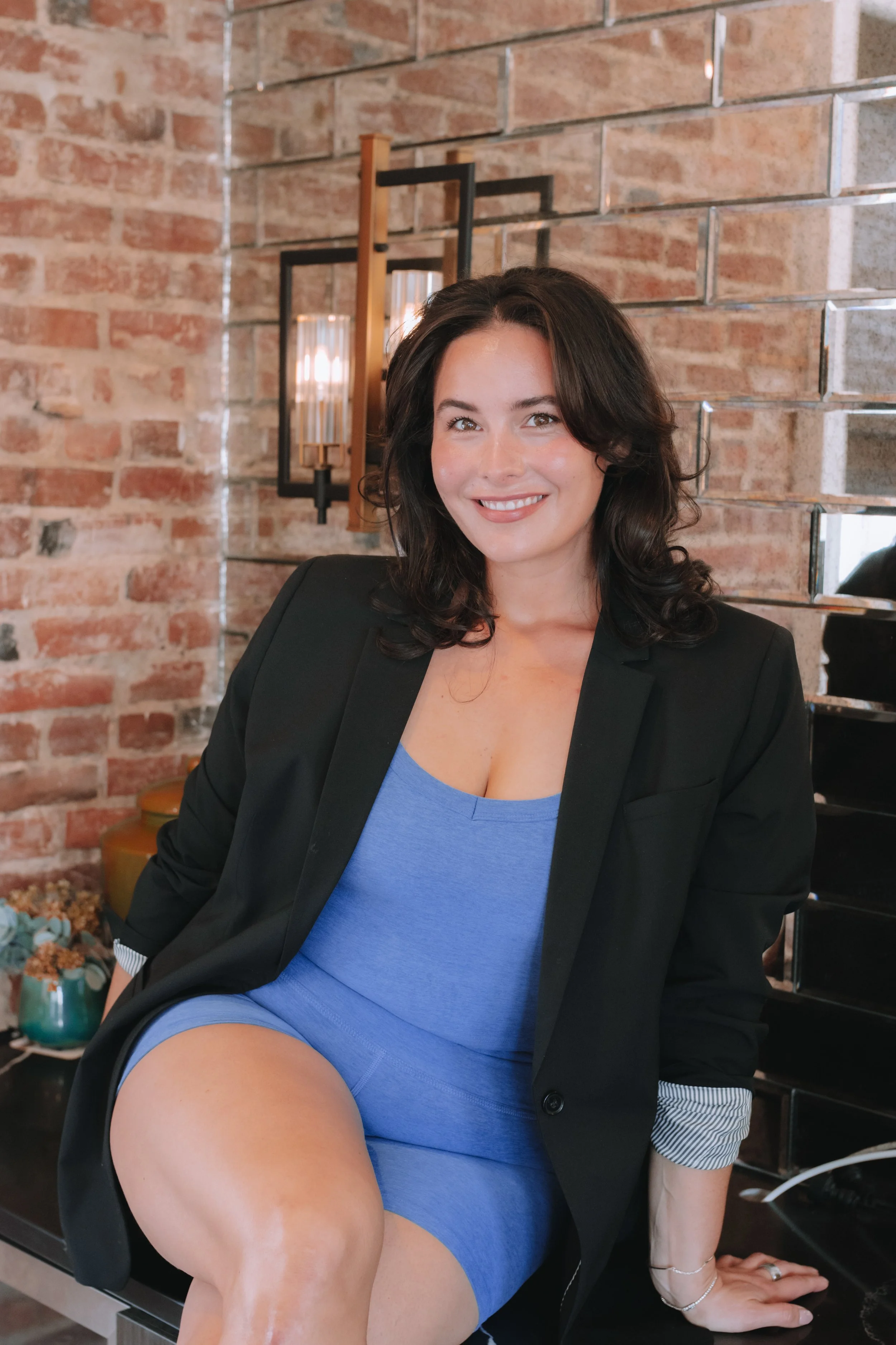 A woman with dark wavy hair and a bright smile, wearing a blue dress and a black blazer, sitting on a table against a brick wall background with wall decor and a mirror.