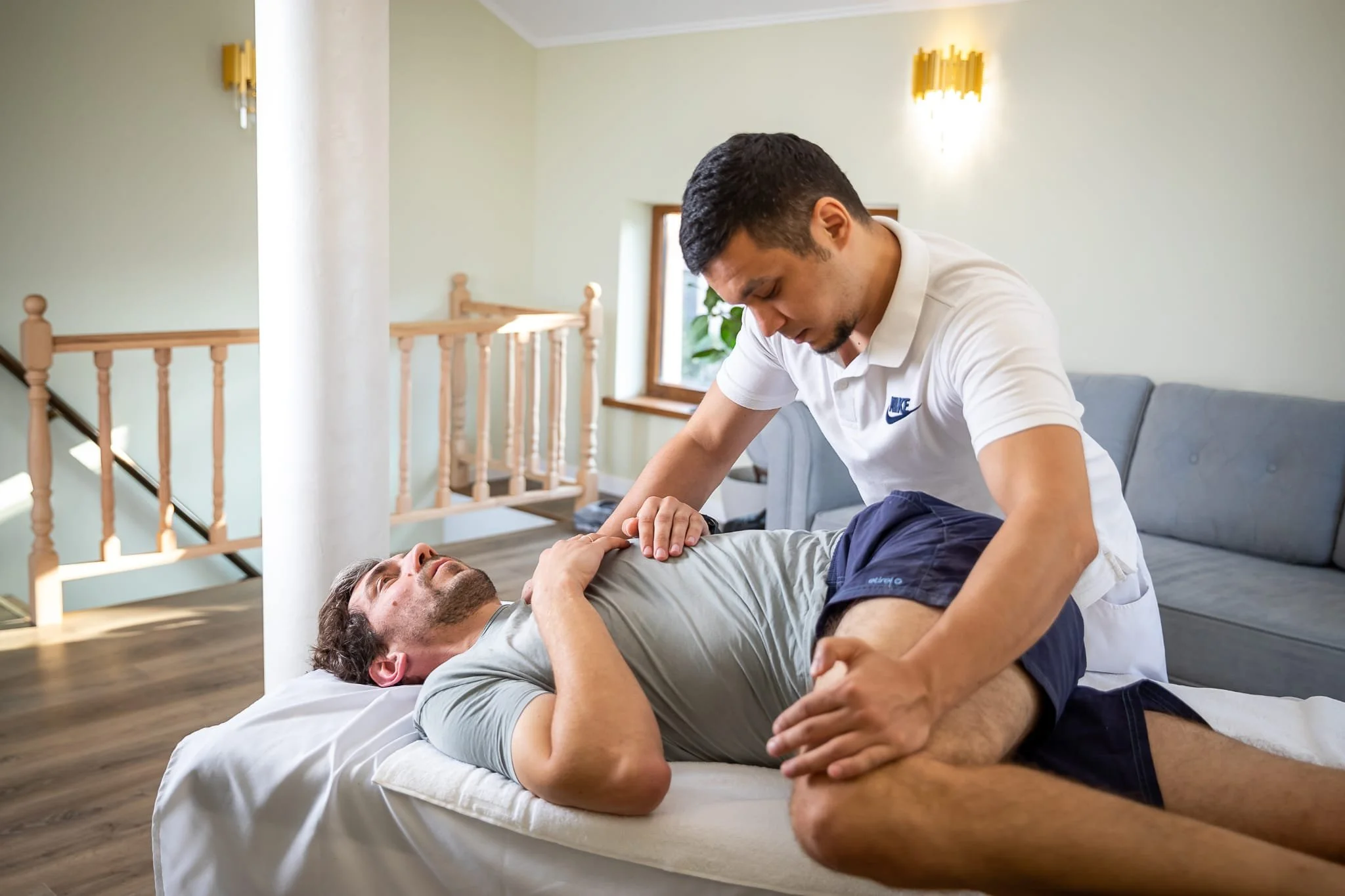 Ein Mann wird von einem Therapeuten im Bett behandelt, vermutlich bei einer physiotherapeutischen Übung oder Massage in einem hellen Raum mit Fenster und Holztreppe im Hintergrund.