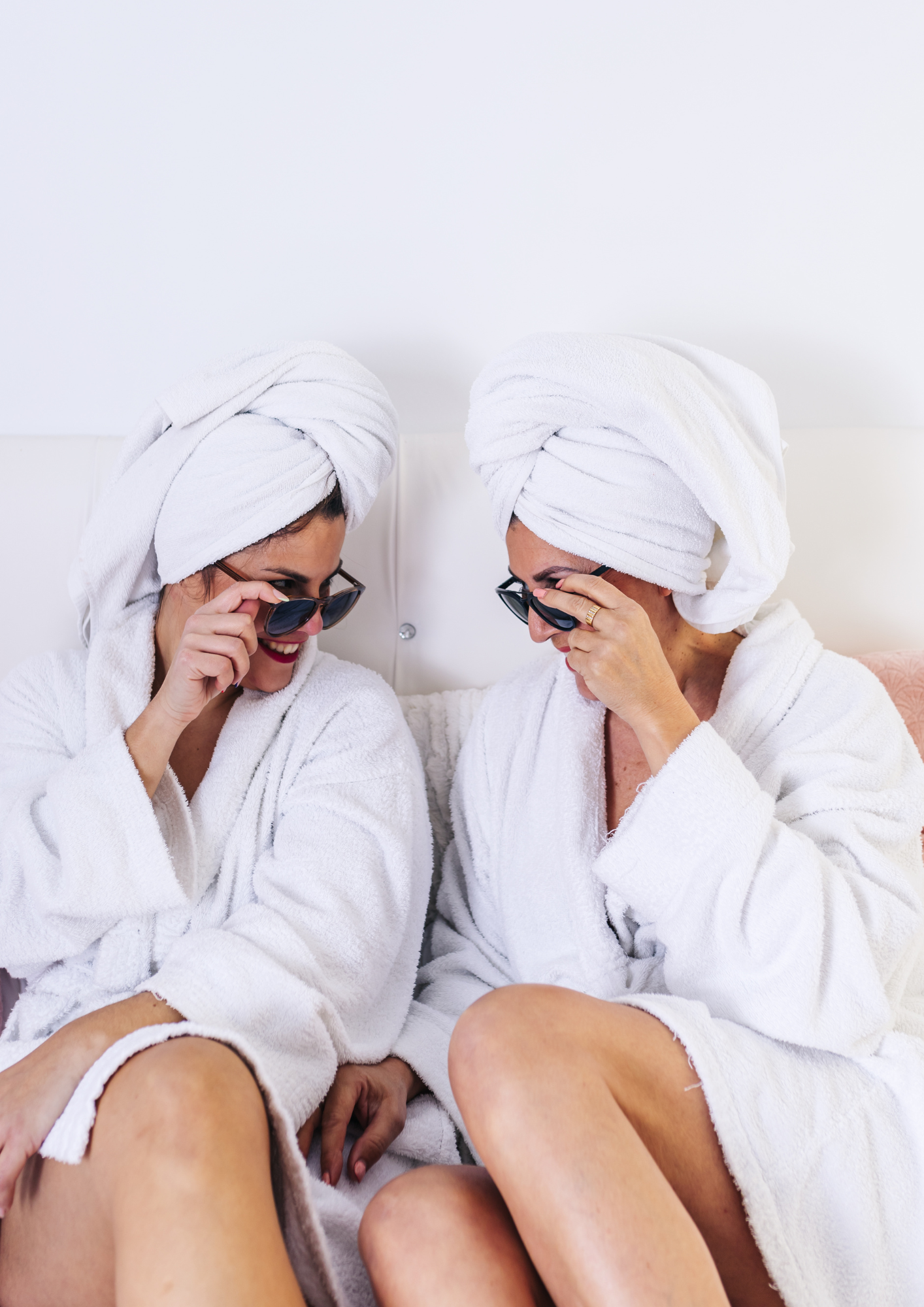 Two women wearing white bathrobes and head towels, sitting close together, smiling, and looking at each other while holding sunglasses in their hands.