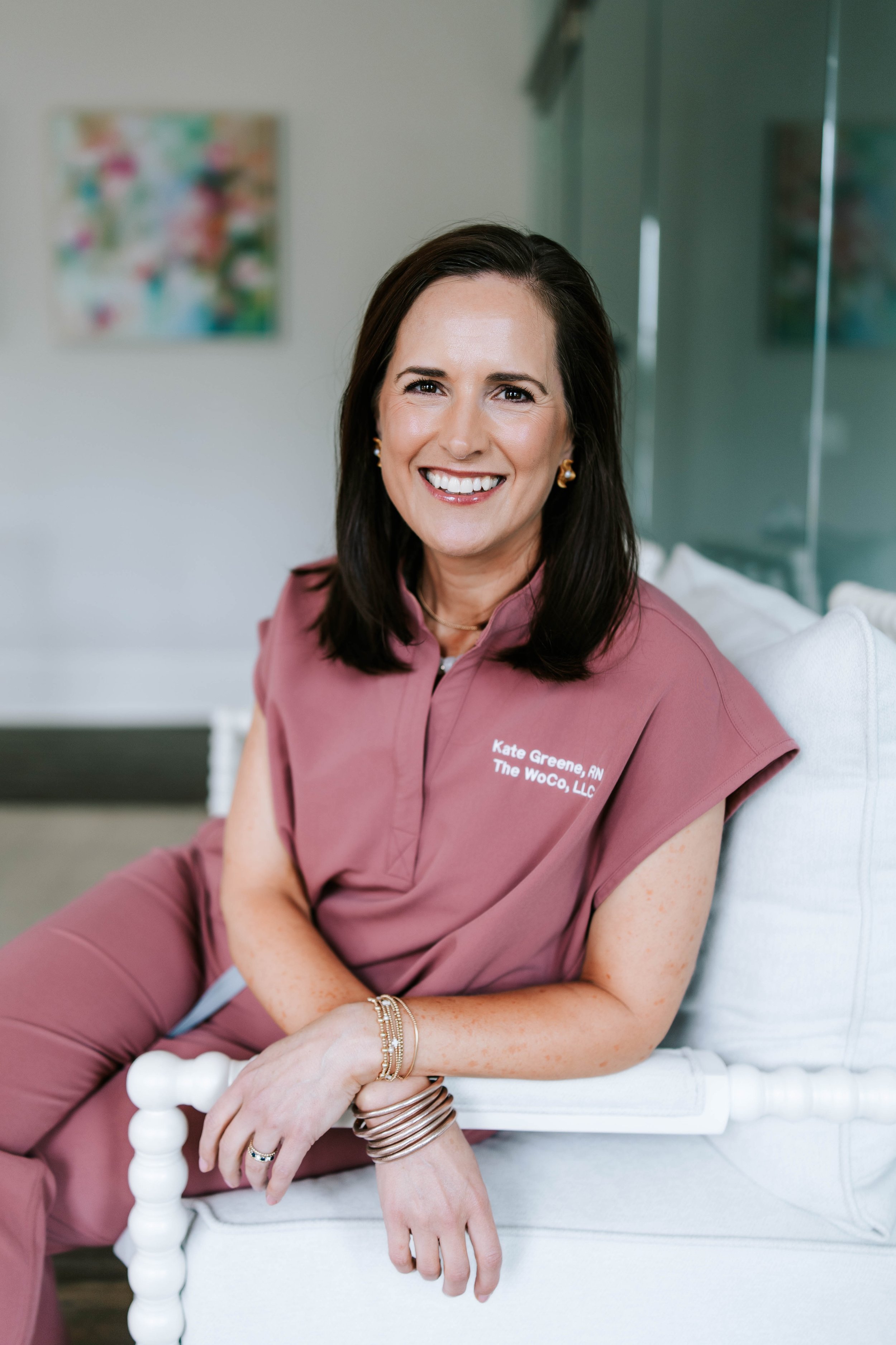 A smiling woman with dark brown hair, wearing pearl earrings and a salmon-colored scrub top with her name and credentials embroidered on it, standing against a plain background. Women's Health Company, Athens, Oconee, Jefferson, Georgia