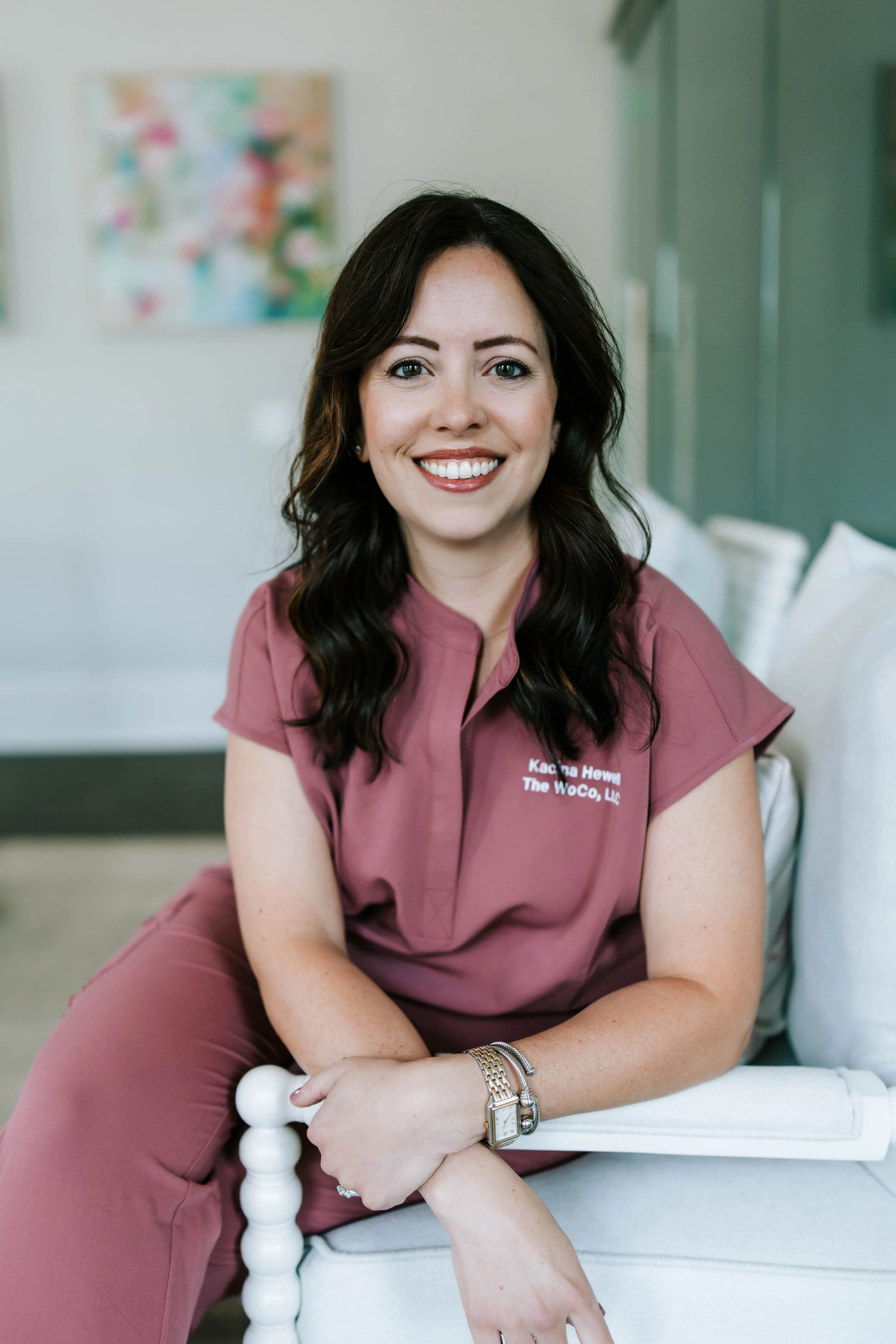 A woman with dark wavy hair, wearing mauve scrubs, sitting on a white bench in a room with artwork on the wall. She is smiling and has a watch and bracelets on her left wrist.