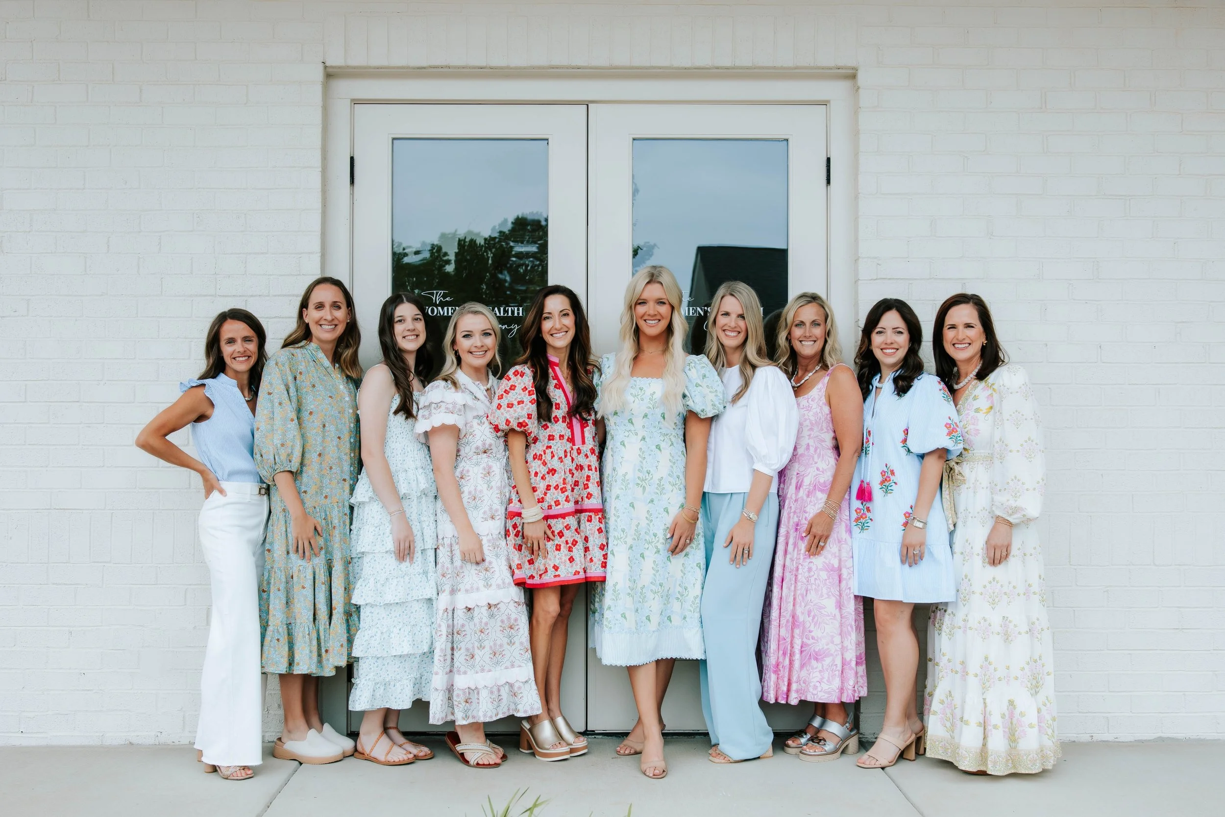 Group of women standing outside in front of a white brick building, smiling and dressed in various colorful dresses.