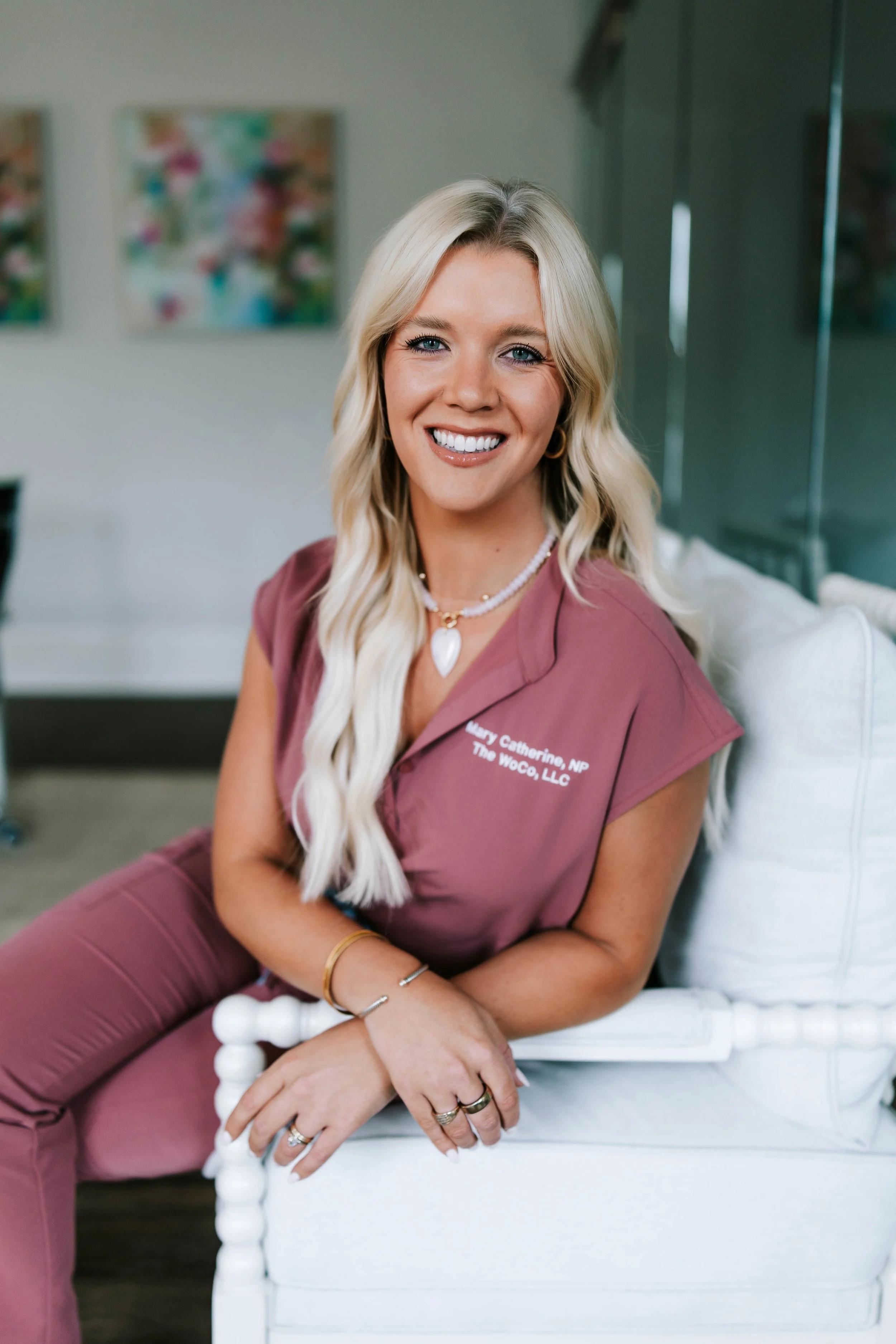 A smiling woman with long blonde hair, wearing a pink medical scrub top and matching bottoms, standing against a white background. Women's Health Company, Athens, Oconee, Jefferson, Georgia