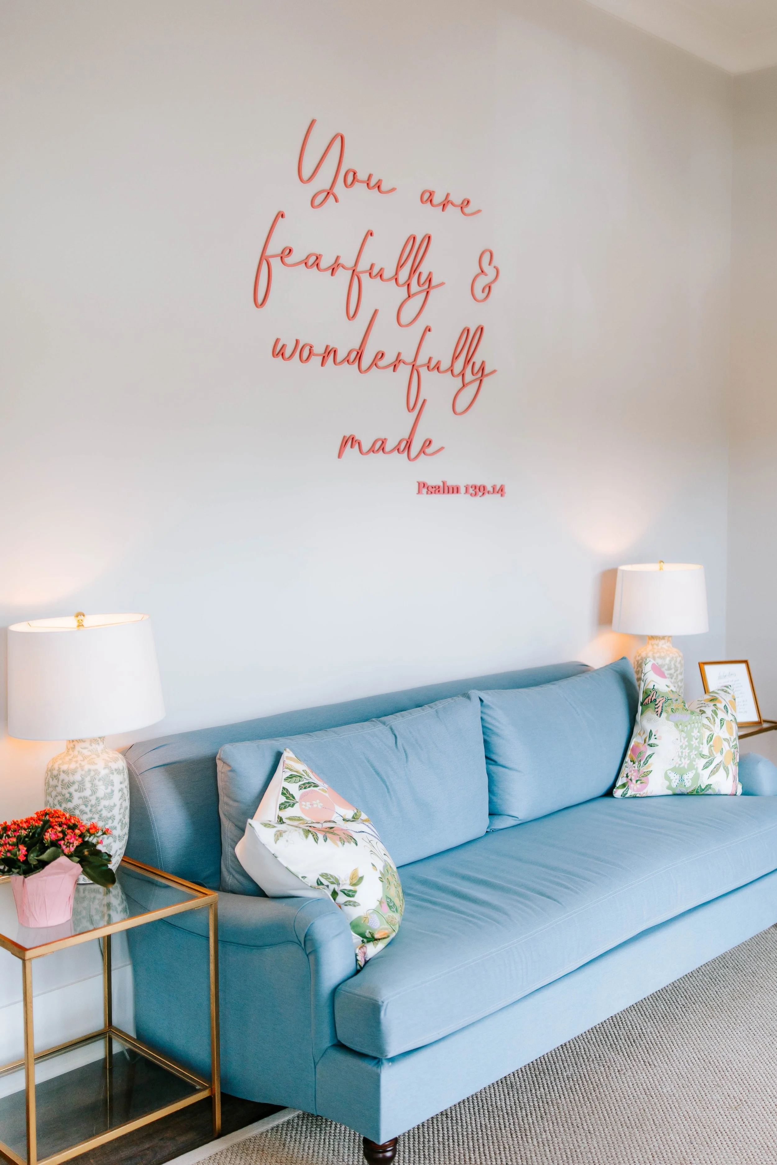 A light blue sofa with floral and pink pillows, flanked by matching lamps on side tables, against a white wall with a pink wall quote quote that says "You are fearfully & wonderfully made" from Psalms 139:14.
