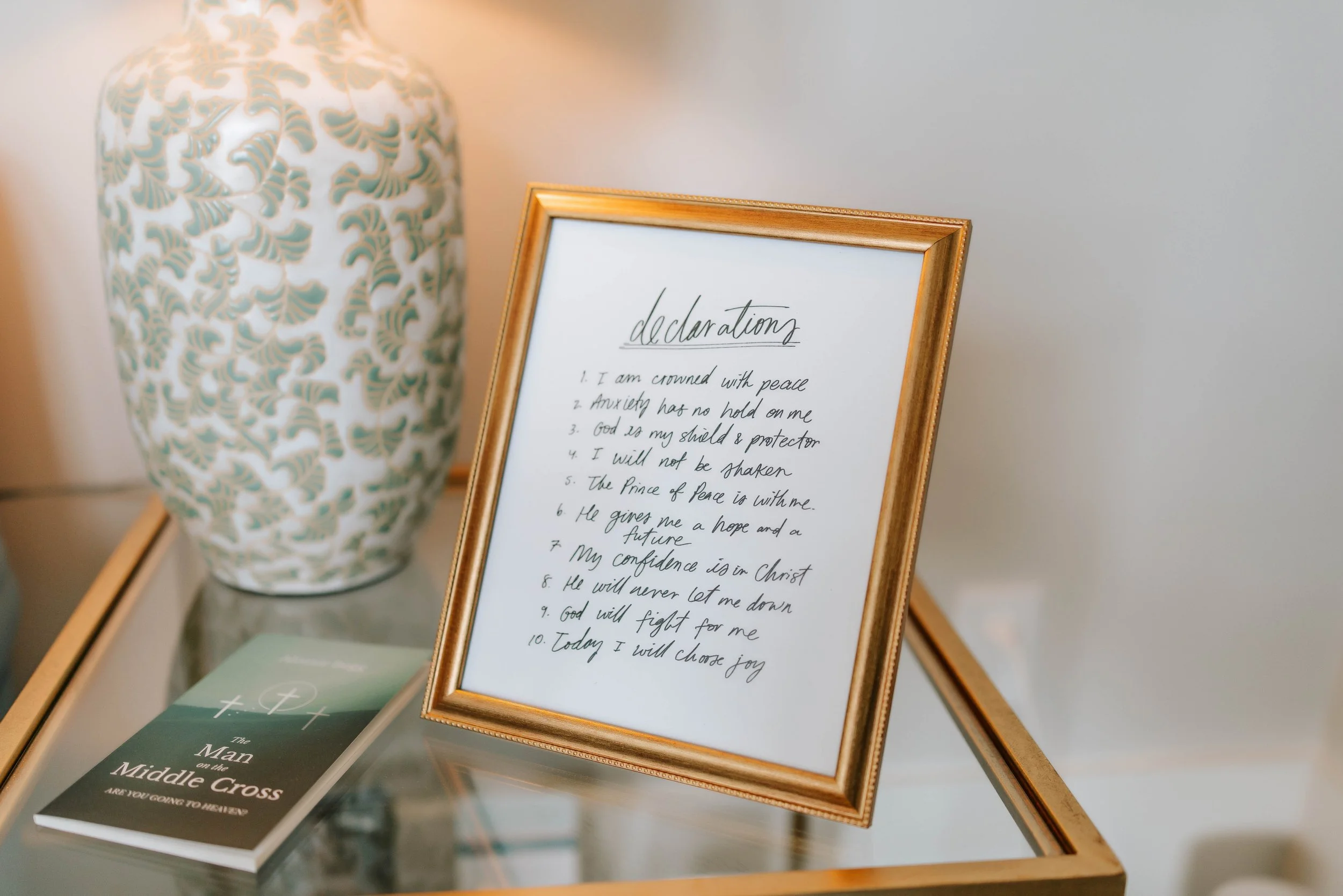 A framed handwritten declaration of faith resting on a surface, with a decorative vase in the background and brochures or pamphlets below.