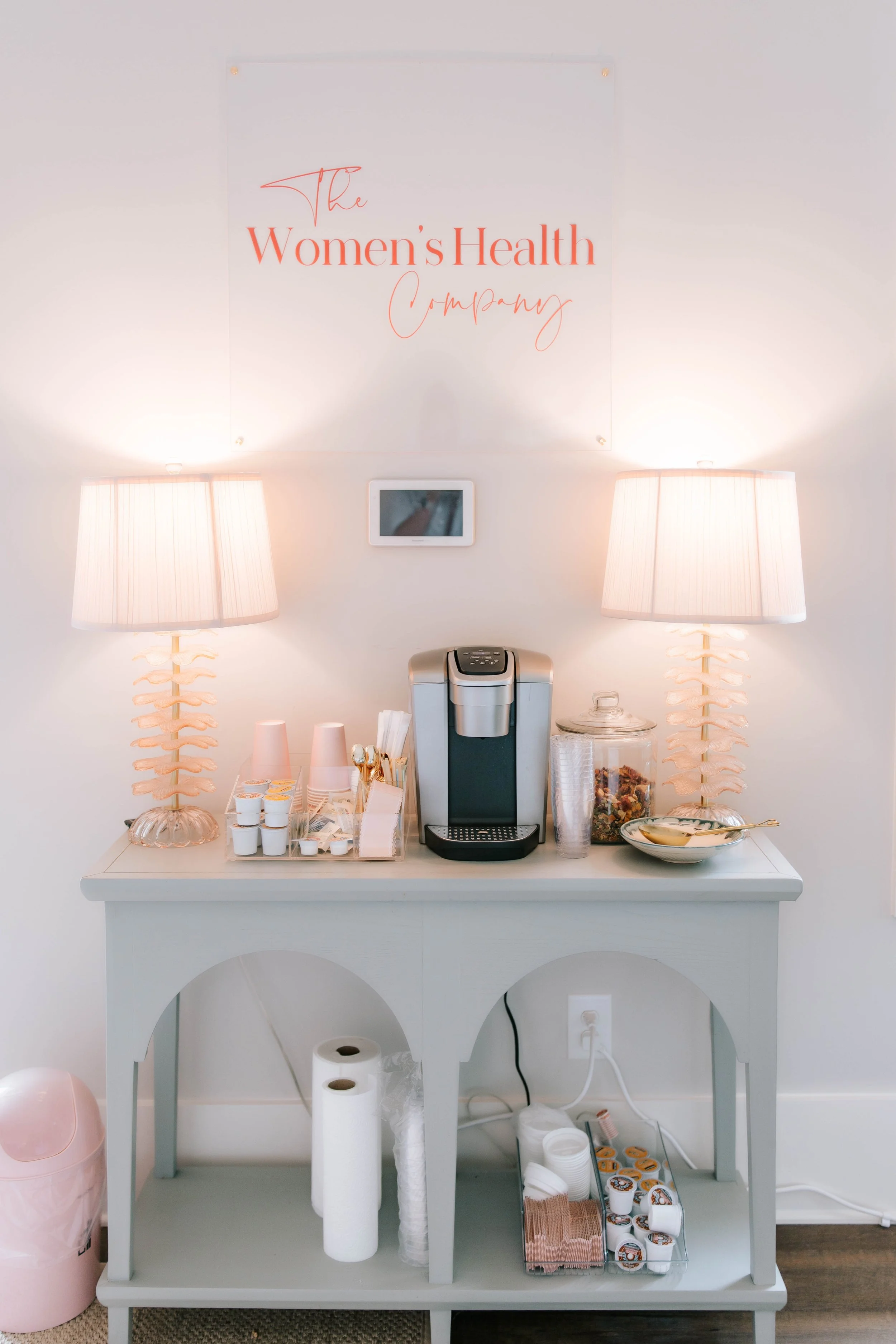 A white table with disposable cups, napkins, and paper towels, a coffee machine, and a glass jar with snacks, flanked by two matching lamps, with a sign above reading 'The Women's Health Company.'