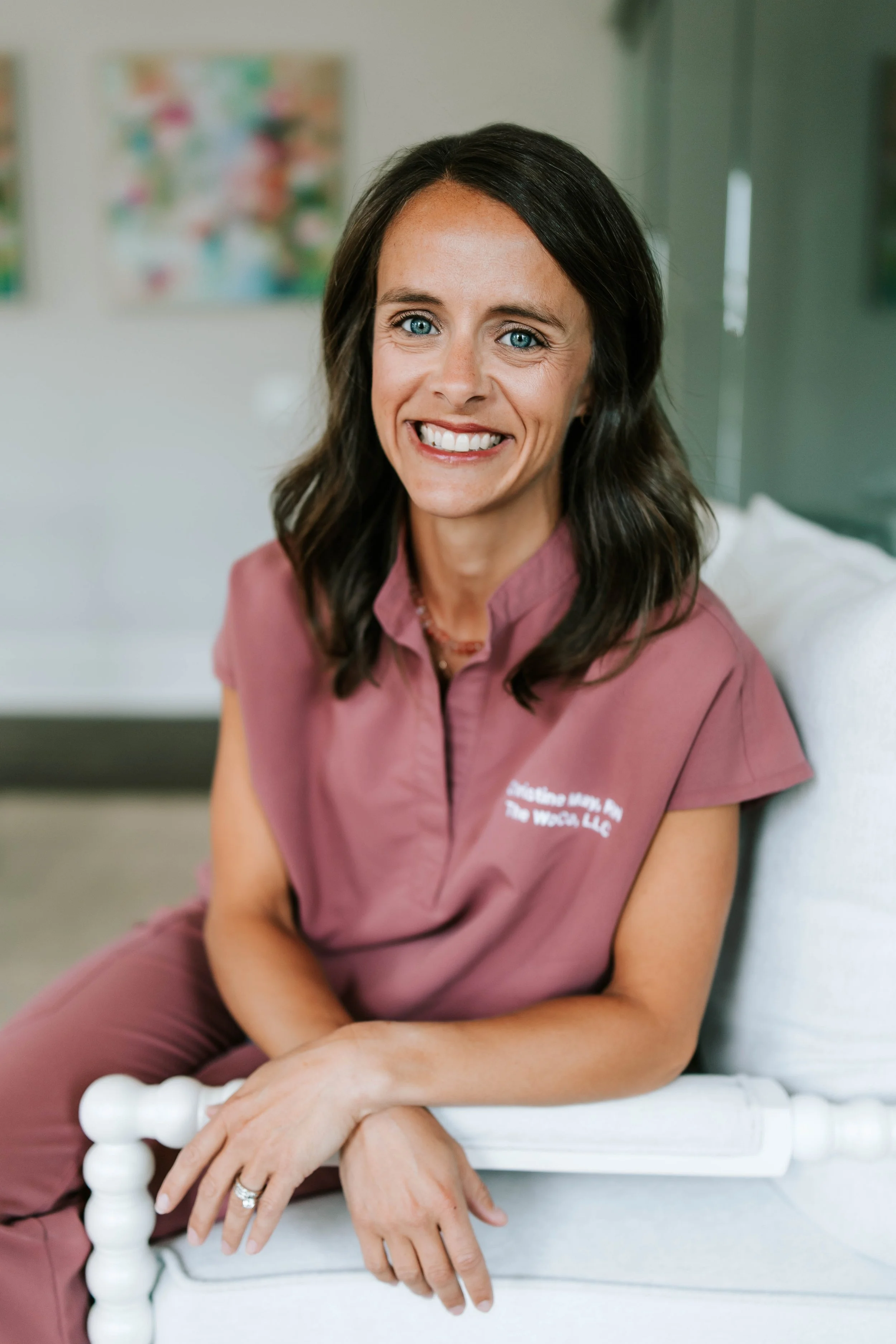 A woman with shoulder-length brown hair and blue eyes smiling, wearing a pink medical uniform with embroidered text, standing with her arms crossed, against a plain white background. Women's Health Company, Athens, Oconee, Jefferson, Georgia