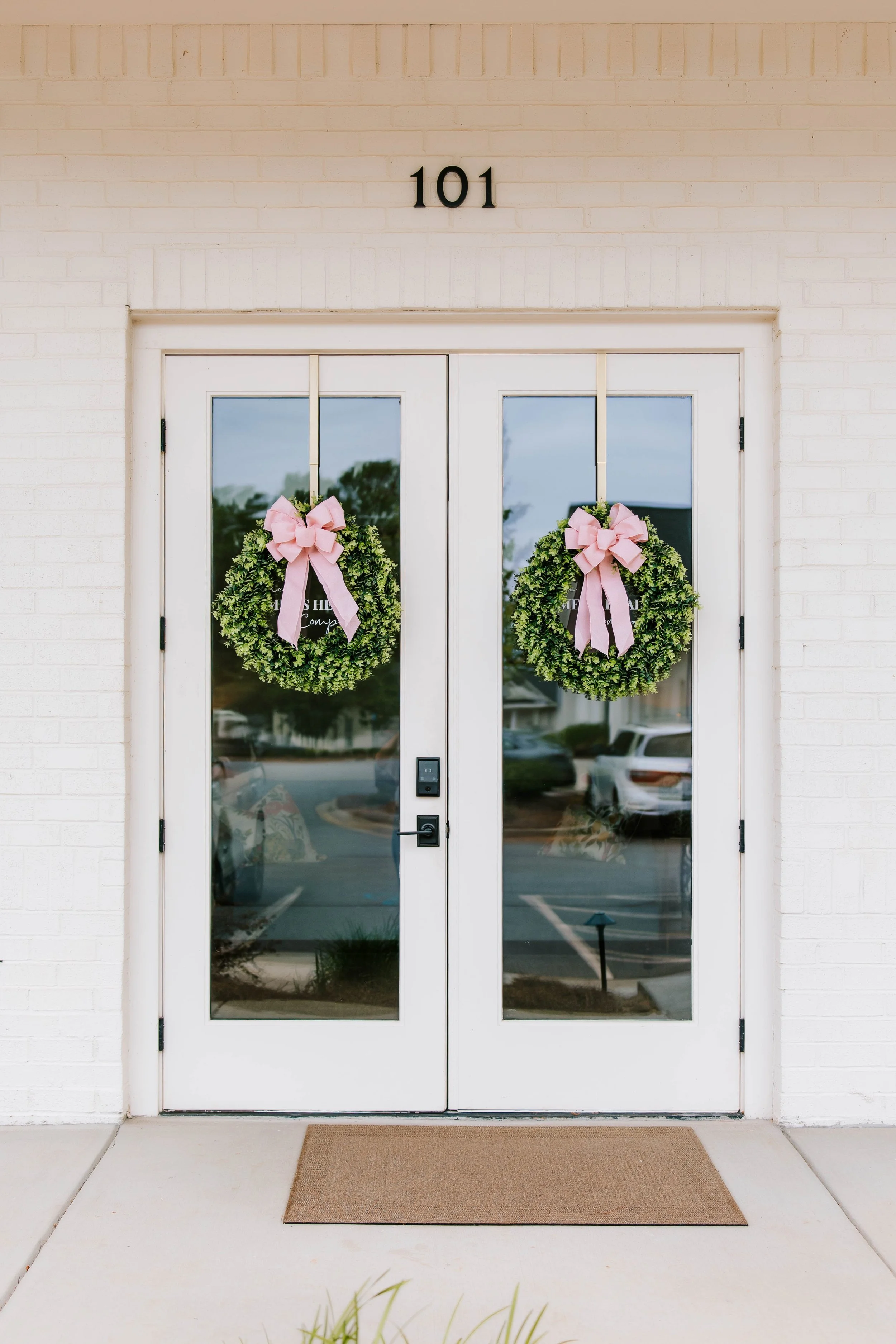 White double glass doors with two matching pink ribbon wreaths hanging on each door, painted brick wall above with the number 101, brown doormat in front, and parking lot visible through the glass.