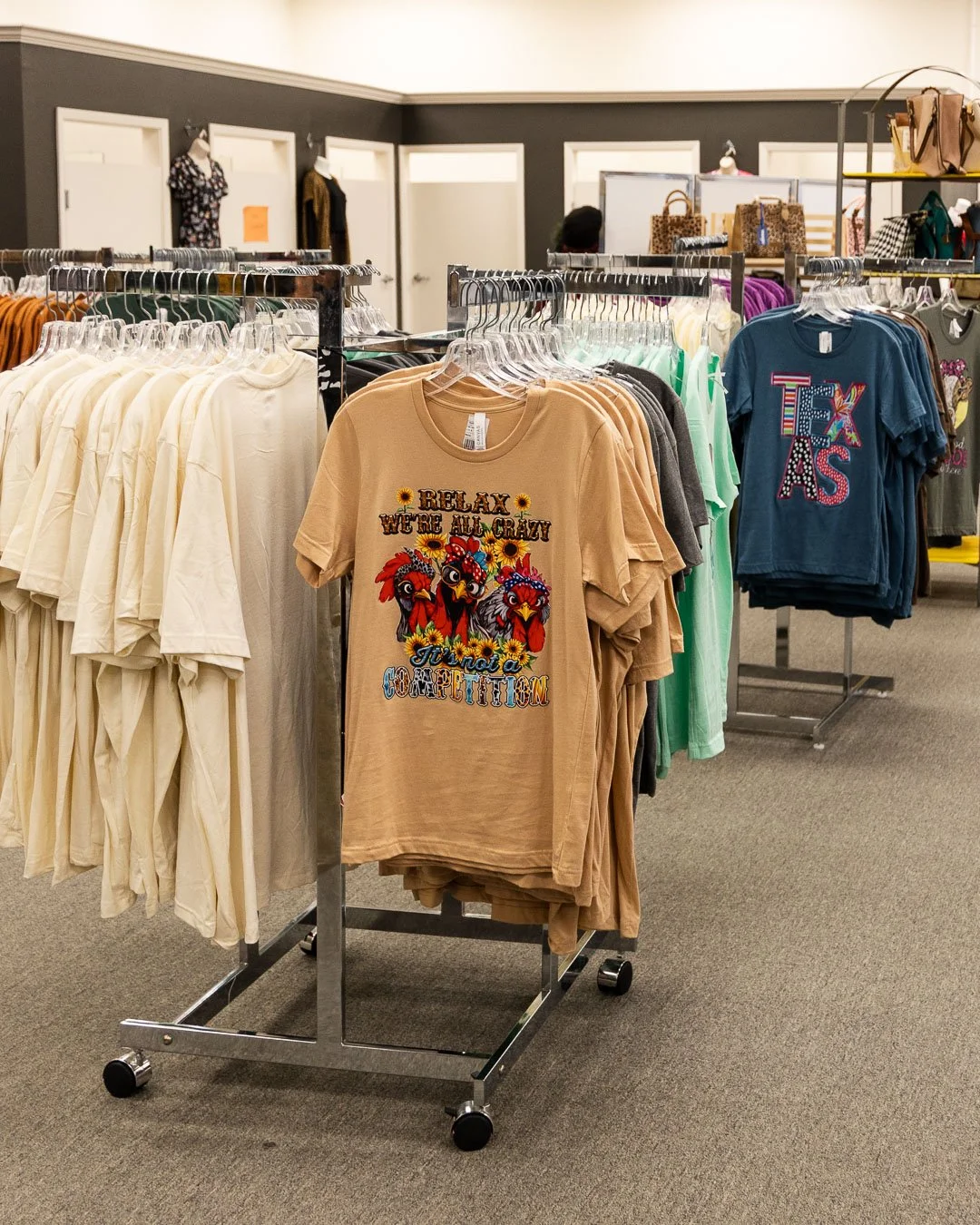 Display of t-shirts on racks in a clothing store. The shirts feature colorful and playful designs, including a reference to Texas.