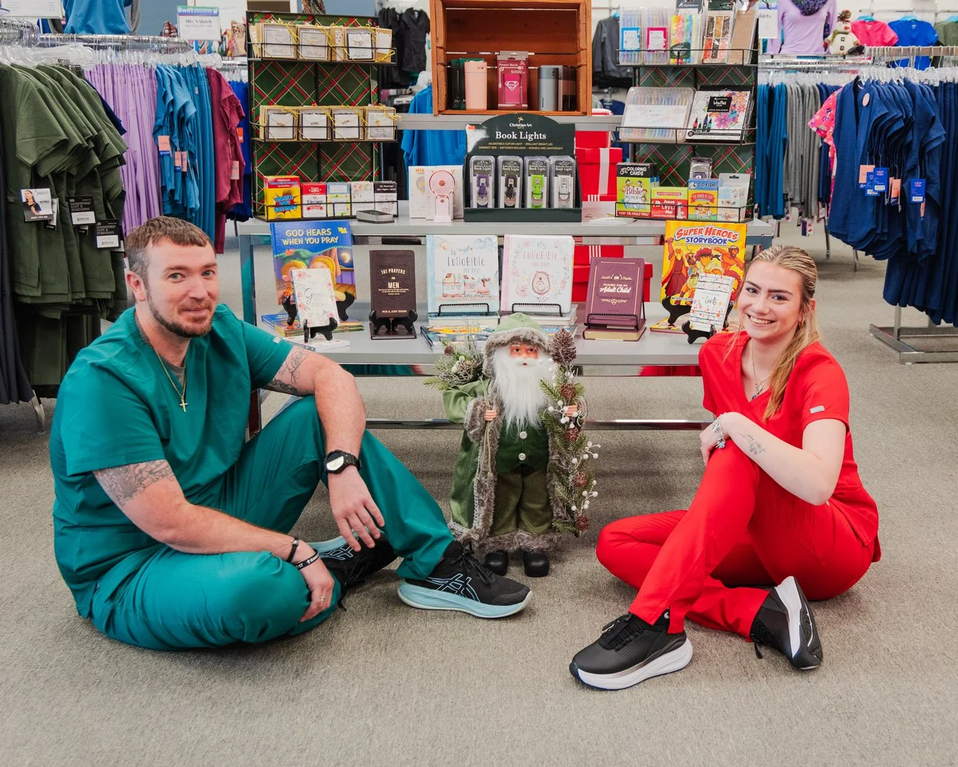 A man and woman sitting on the floor of a store, posing next to a Santa Claus figurine and holiday-themed displays. The store has clothing racks and shelves in the background.
