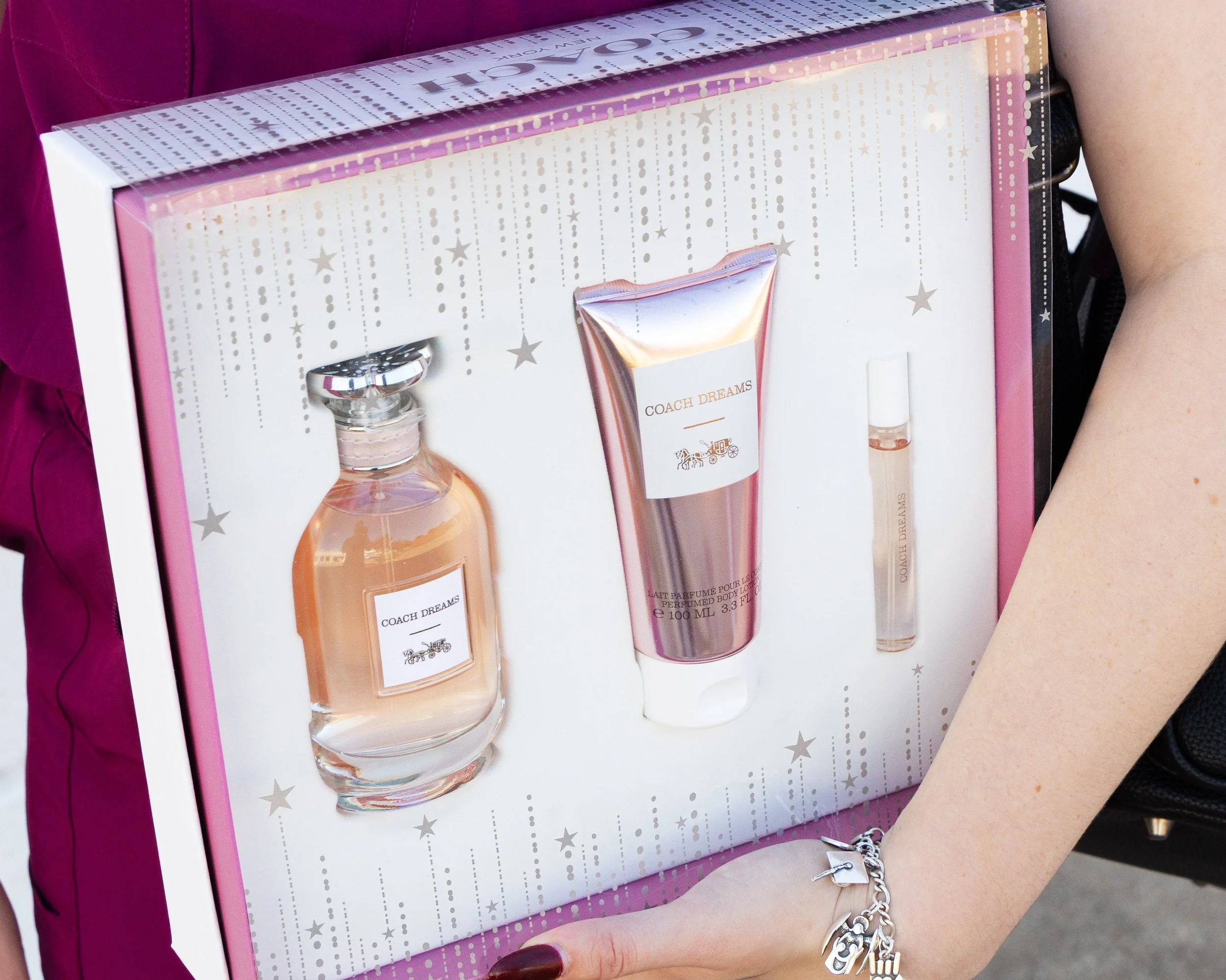 A person holding a Coach Dreams perfume gift set with three items: a perfume bottle, a body lotion tube, and a rollerball perfume, all in a festive box with star and dotted line designs.