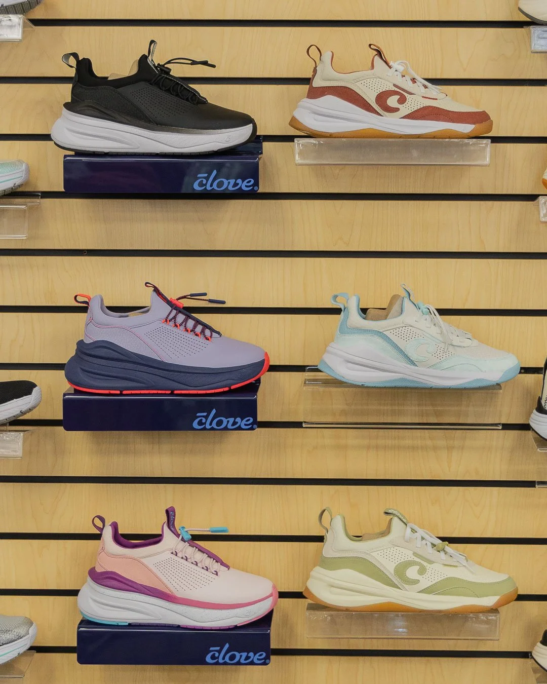Display of six pairs of sneakers on wooden shelves in a shoe store. The sneakers are in various colors including black, cream, lavender, pink, and green, with some placed on boxes labeled 'clove'.