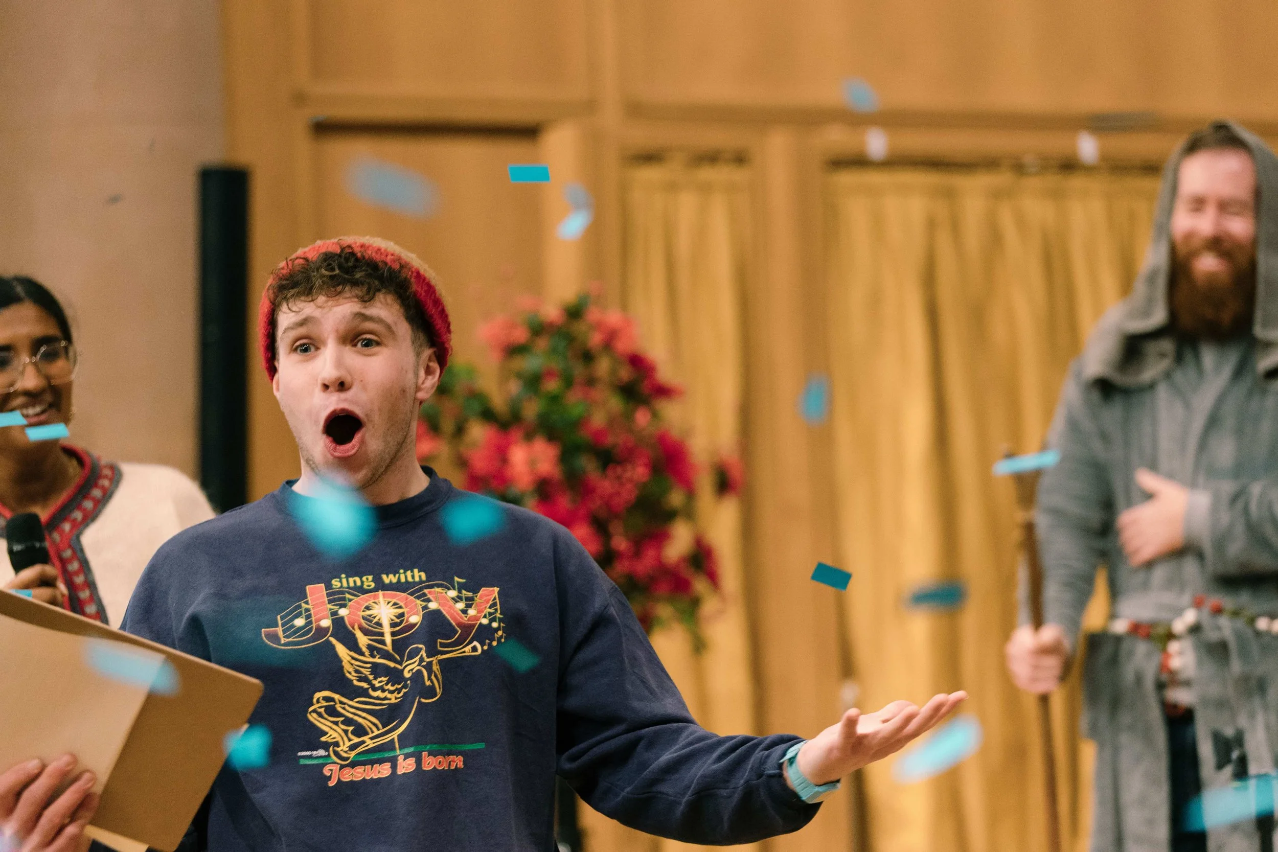 A young man with a surprised expression, wearing a red knit hat and a navy blue sweatshirt with a Christmas-themed design, is celebrating with blue confetti falling around him. A woman with glasses and a microphone is on the left, and a bearded man with a gray hoodie is on the right, all in a room with wooden walls and pink flowers in the background.