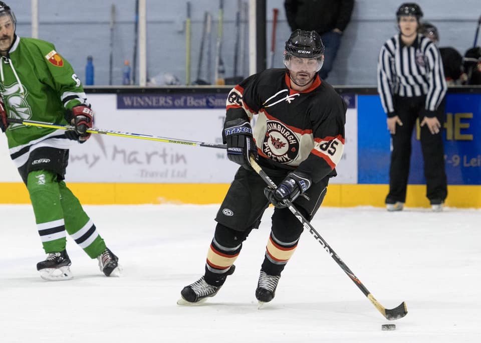 Hockey player in black and orange jersey with a logo, controlling the puck on the ice rink during a game, with a referee and another player in green in the background.