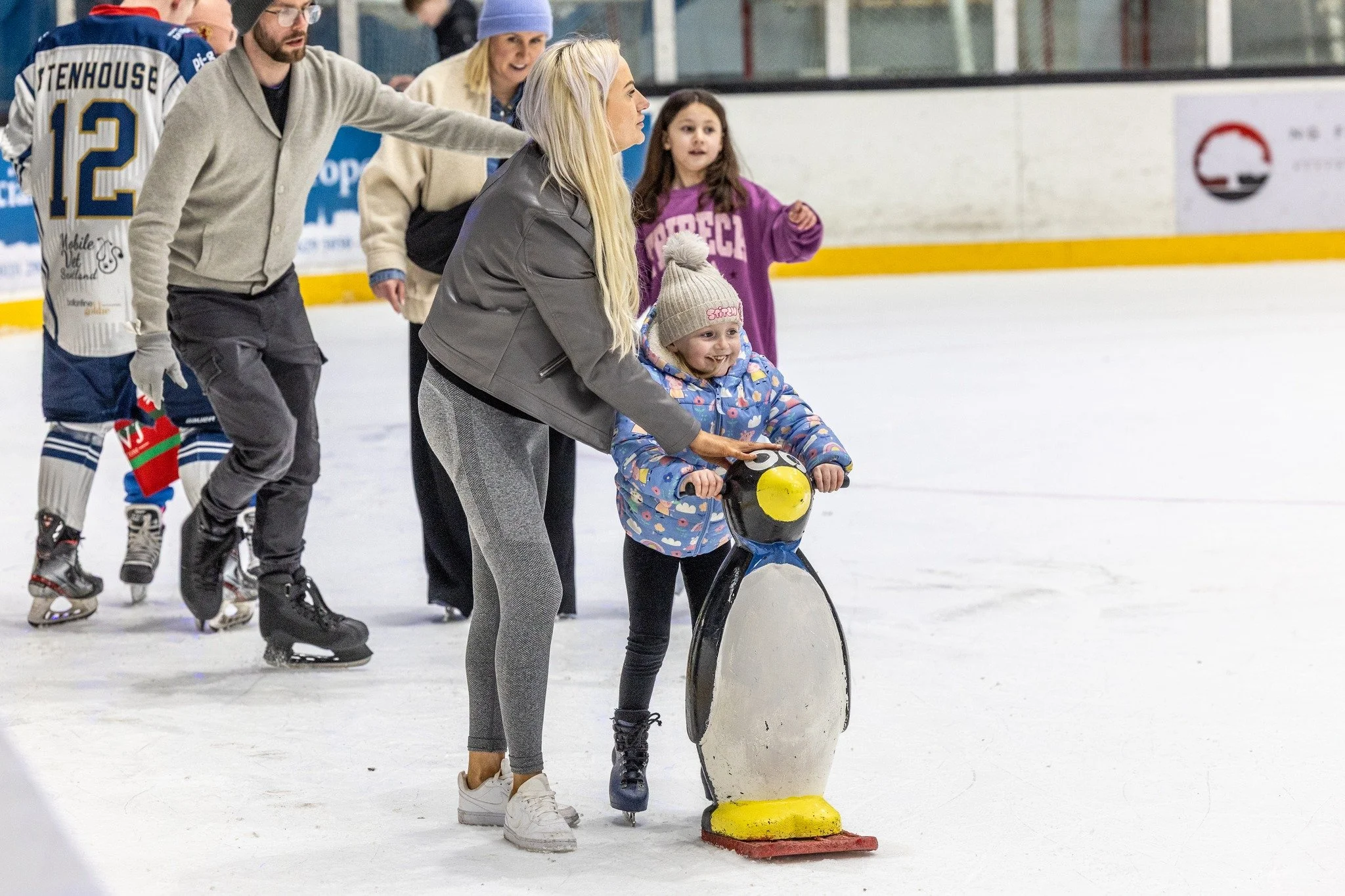 A group of people, including a young girl, on an ice rink. The girl is holding onto a penguin-shaped skating aid, and an adult woman is helping her. Other skaters are visible in the background.