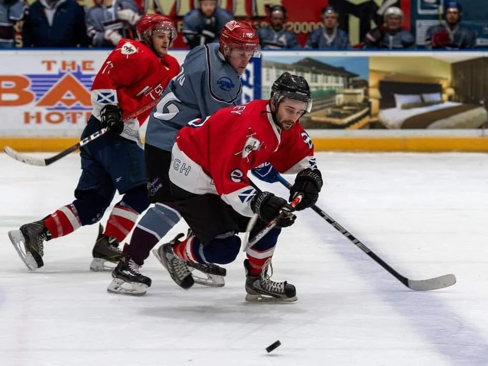 Three hockey players are chasing the puck on the ice rink. One player in a red and white jersey is in front, reaching for the puck with his stick. Two players in red or blue jerseys follow behind him. Spectators watch from the background.