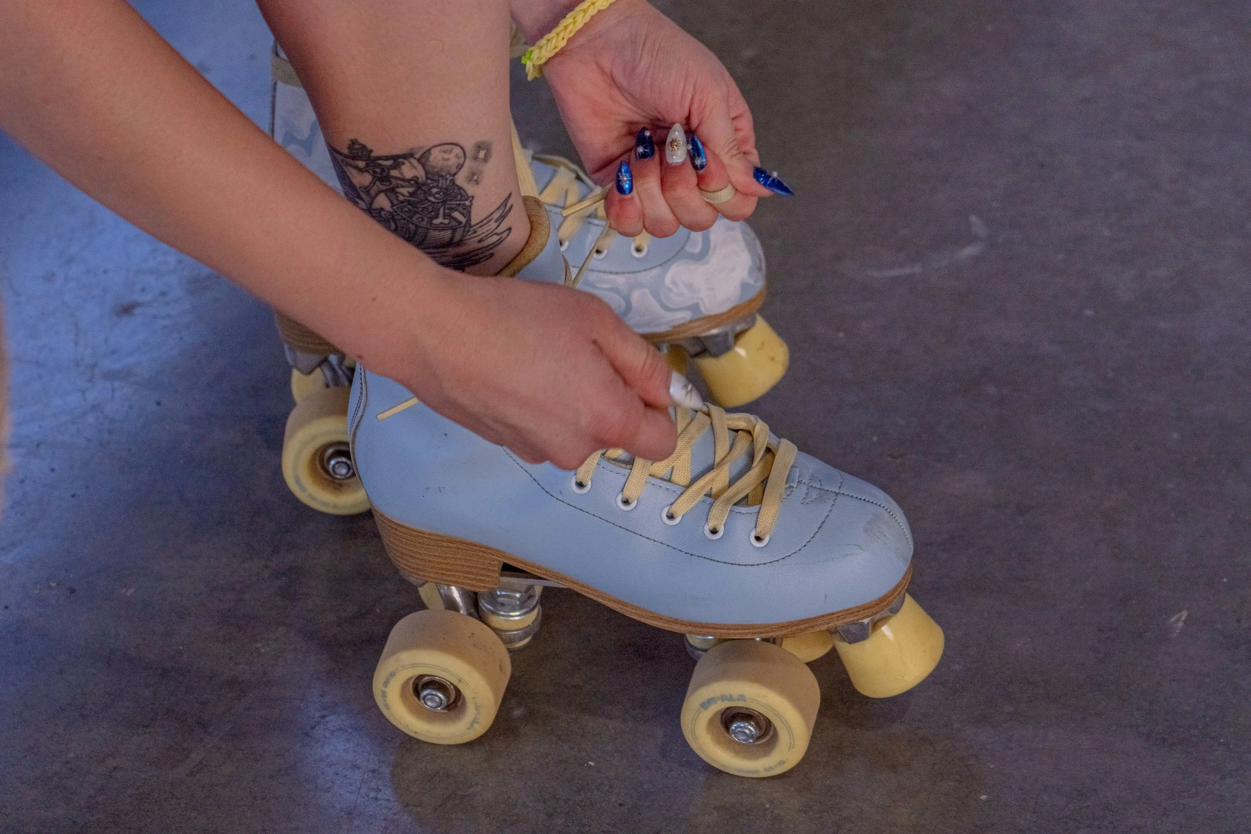Person tying the laces of light blue roller skates with yellow wheels, standing on a gray concrete floor. The person has a tattoo on their arm and painted nails.