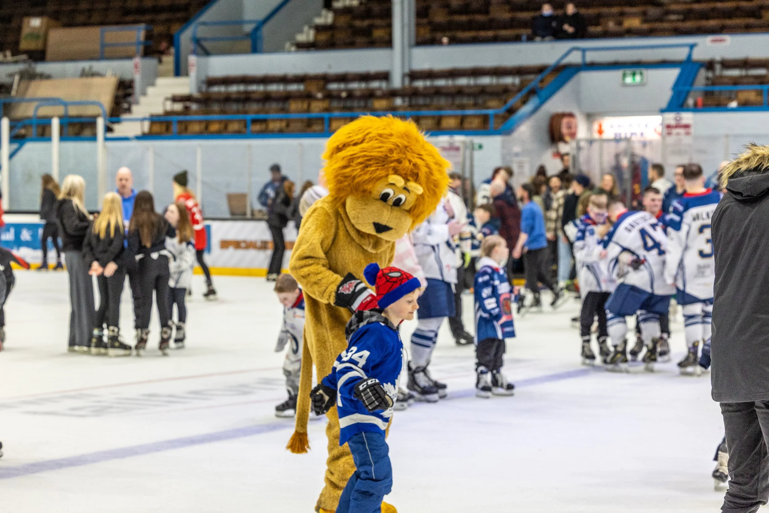 A person dressed in a lion mascot costume interacting with a young boy wearing a blue hockey uniform and a red and blue spiderman hat at an ice hockey rink filled with players and spectators.