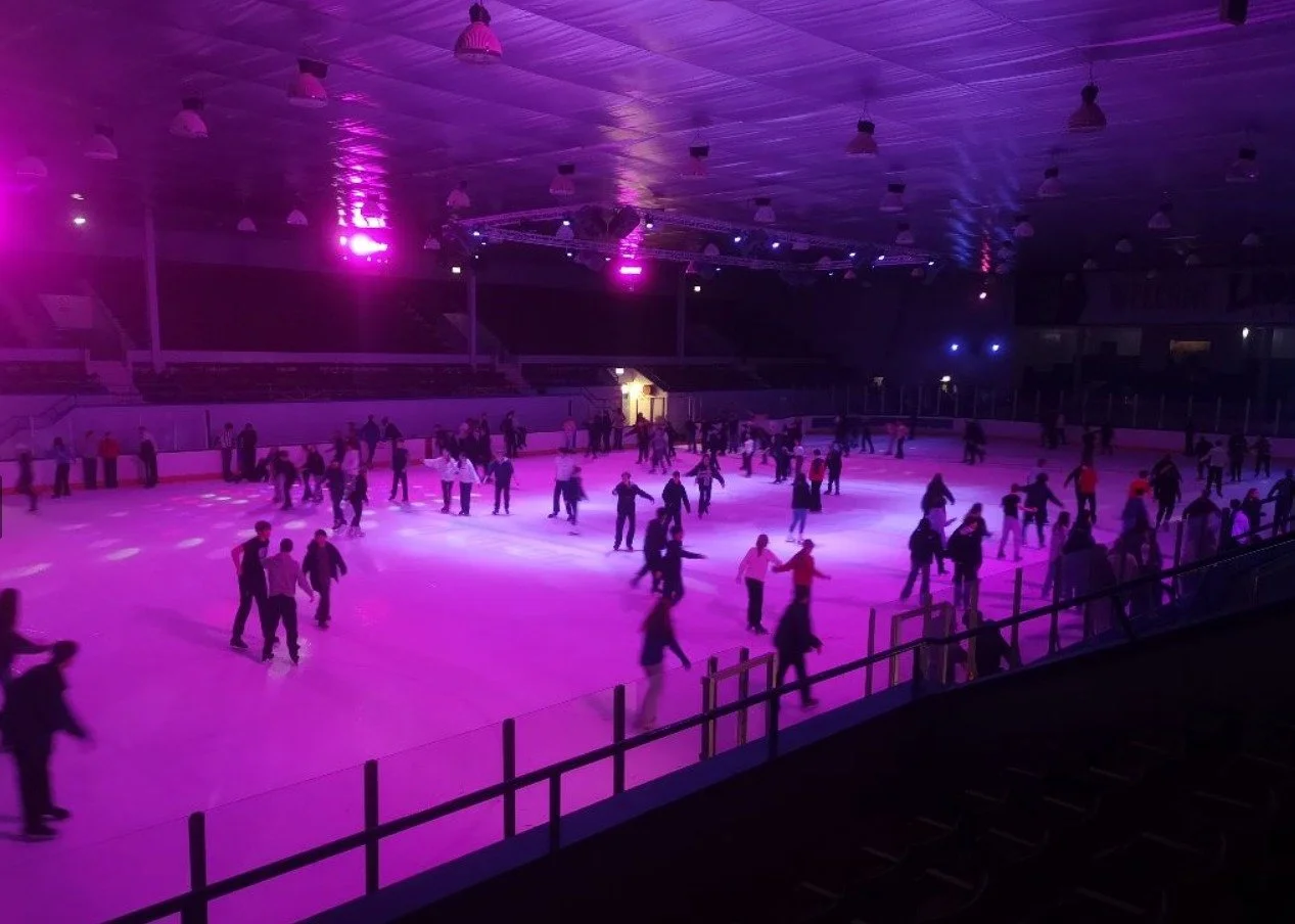 People ice skating at an indoor rink illuminated with purple and pink lights.