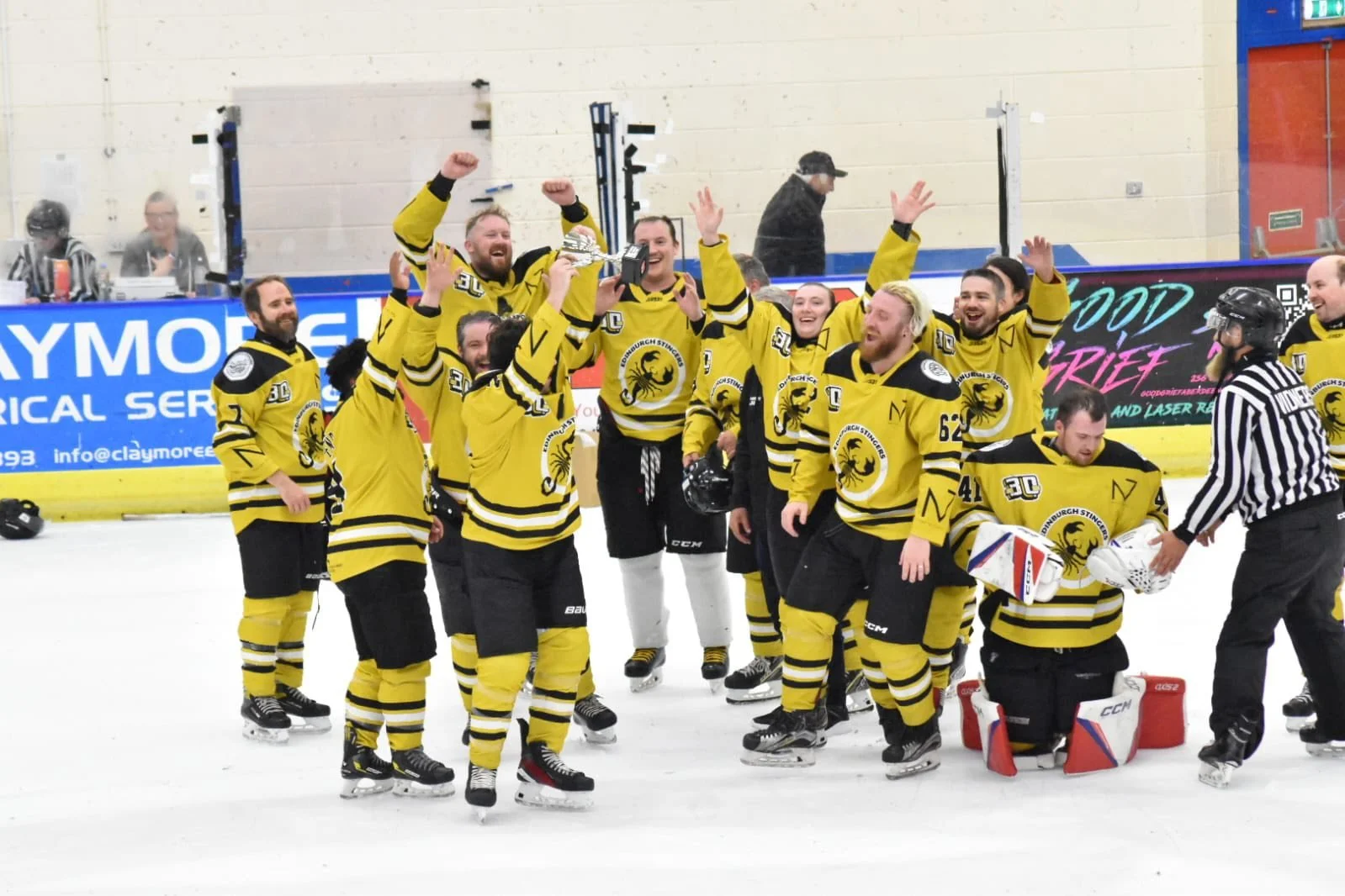 Hockey team in yellow jerseys celebrating on ice rink, with trophy raised, crowd watching in background.