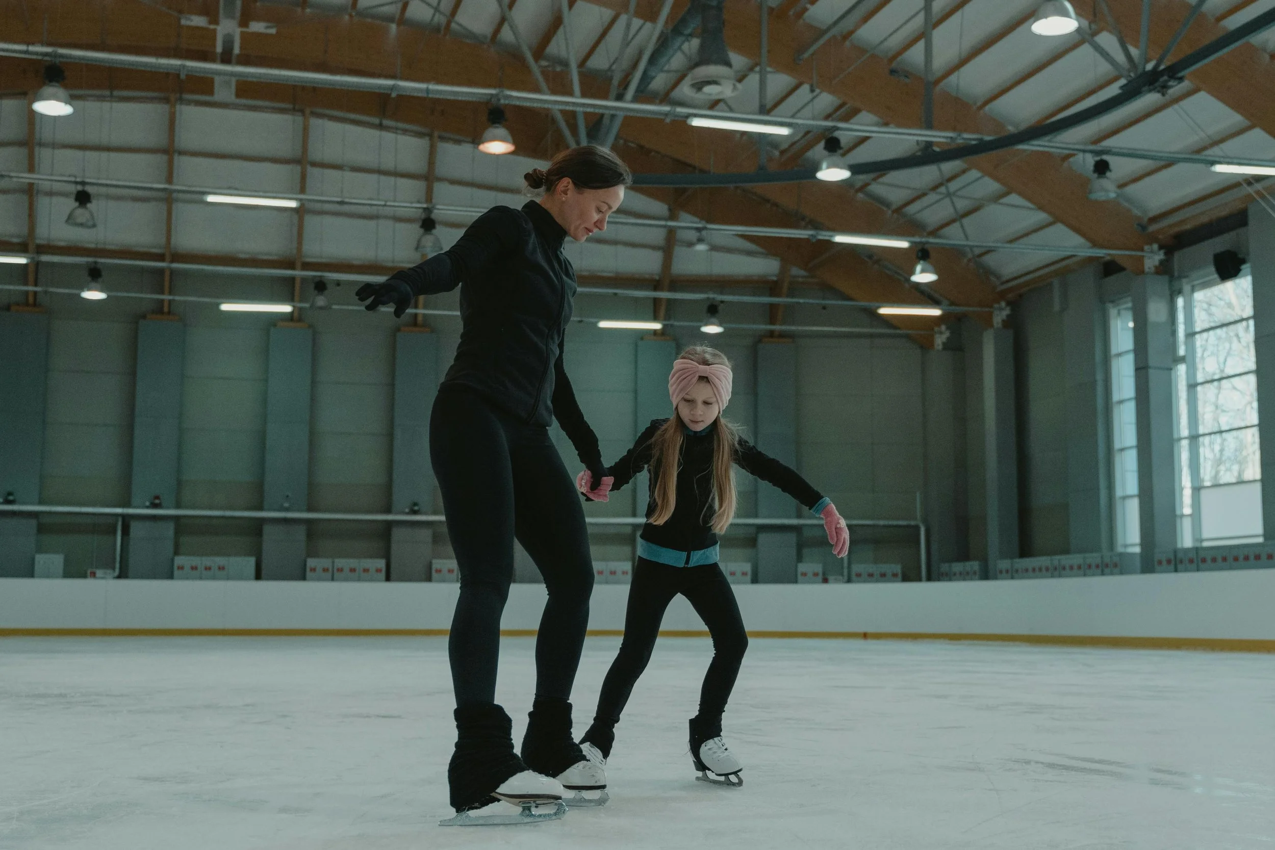 A woman and young girl figure skating on an indoor ice rink, with the woman teaching or assisting the girl.