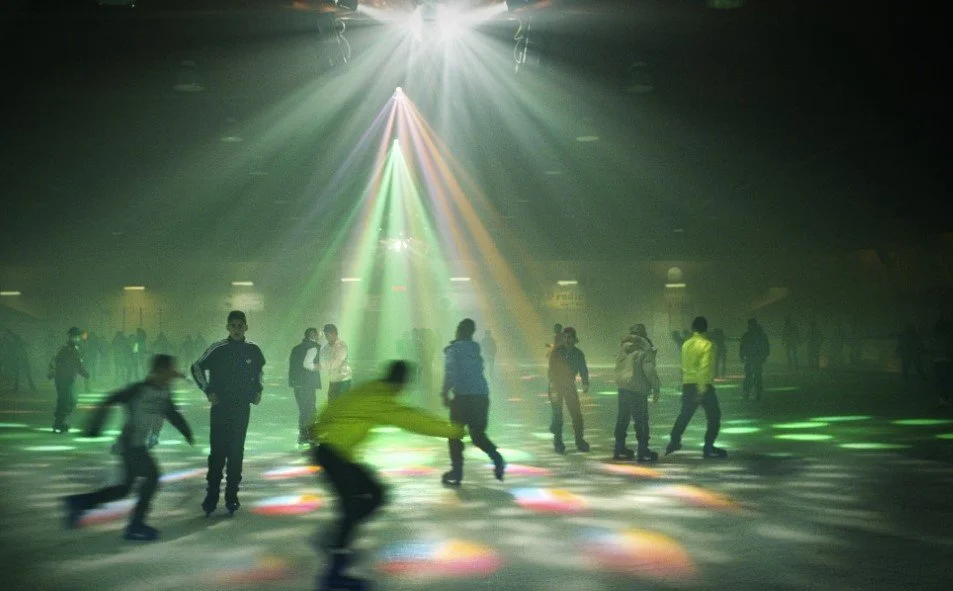 People ice skating at an indoor rink with colorful lights and a bright spotlight overhead.
