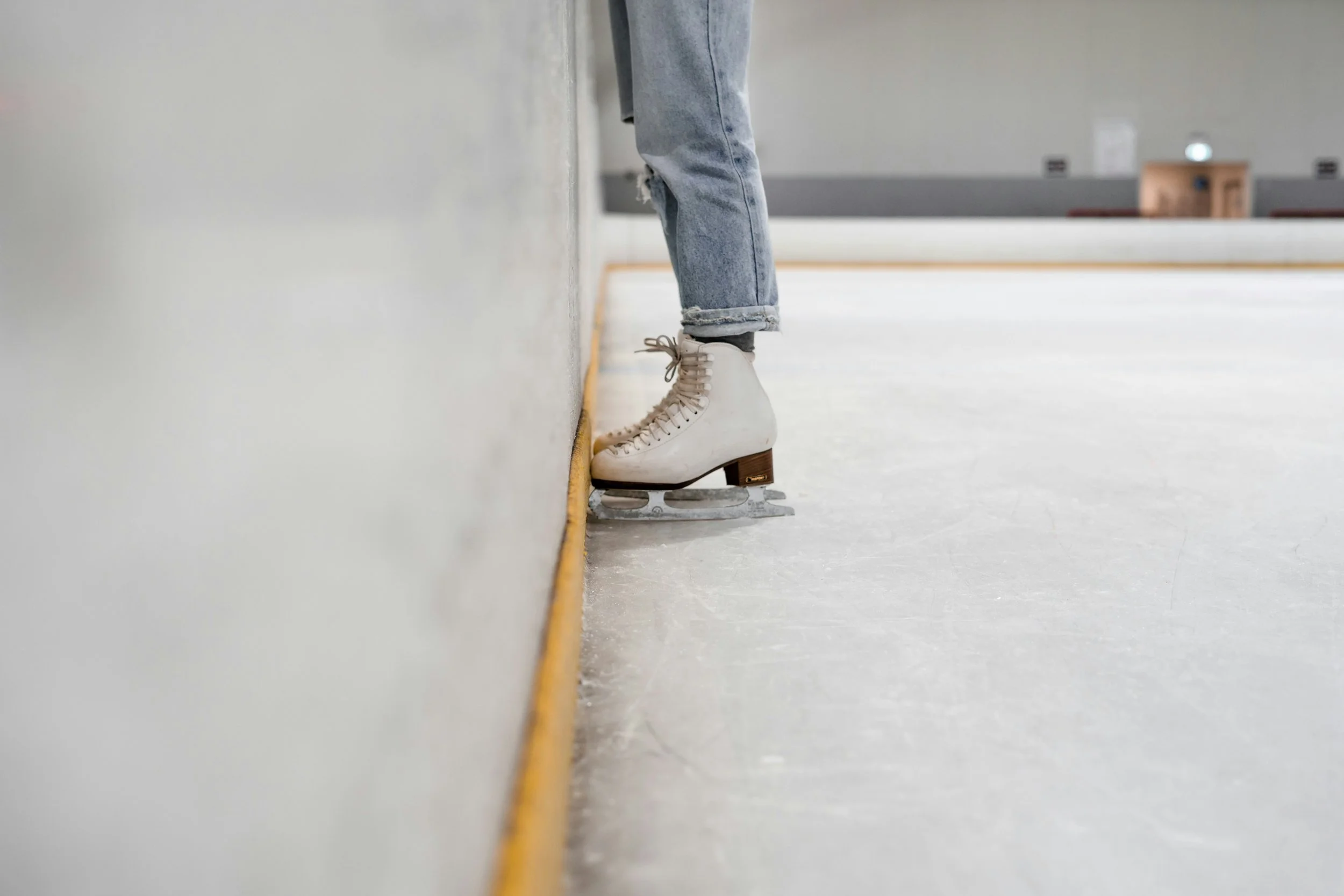 Person wearing white ice skates and ripped jeans standing at the edge of an ice skating rink.