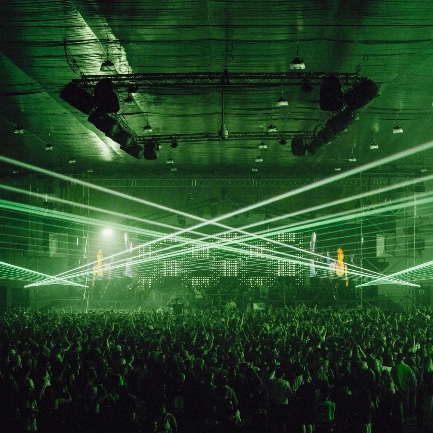 A large indoor concert or electronic music event with a crowd, green laser lights, and a LED screen.