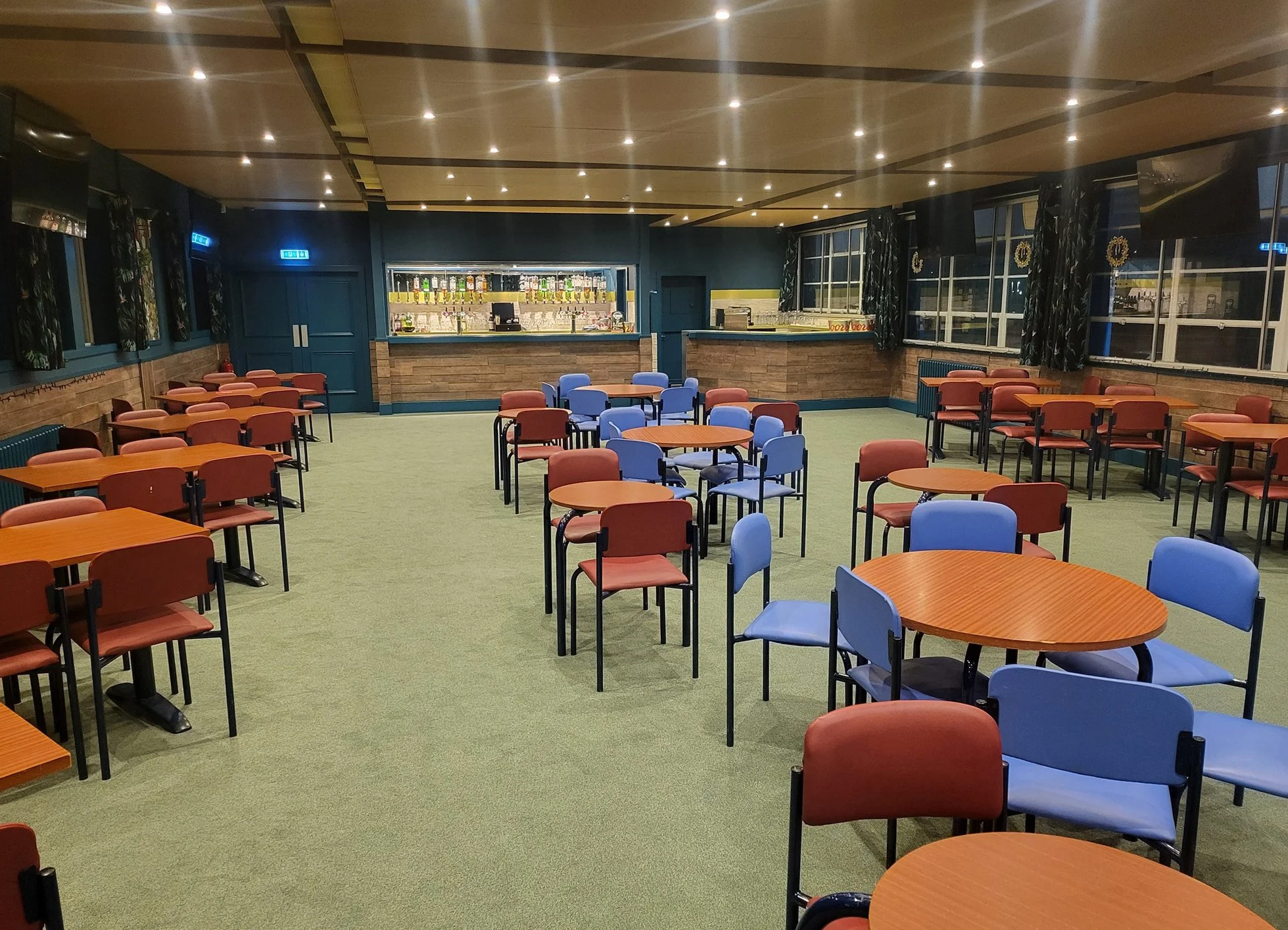 Empty restaurant or bar room with round tables, chairs in red and blue, a bar in the background, and windows along the side walls.