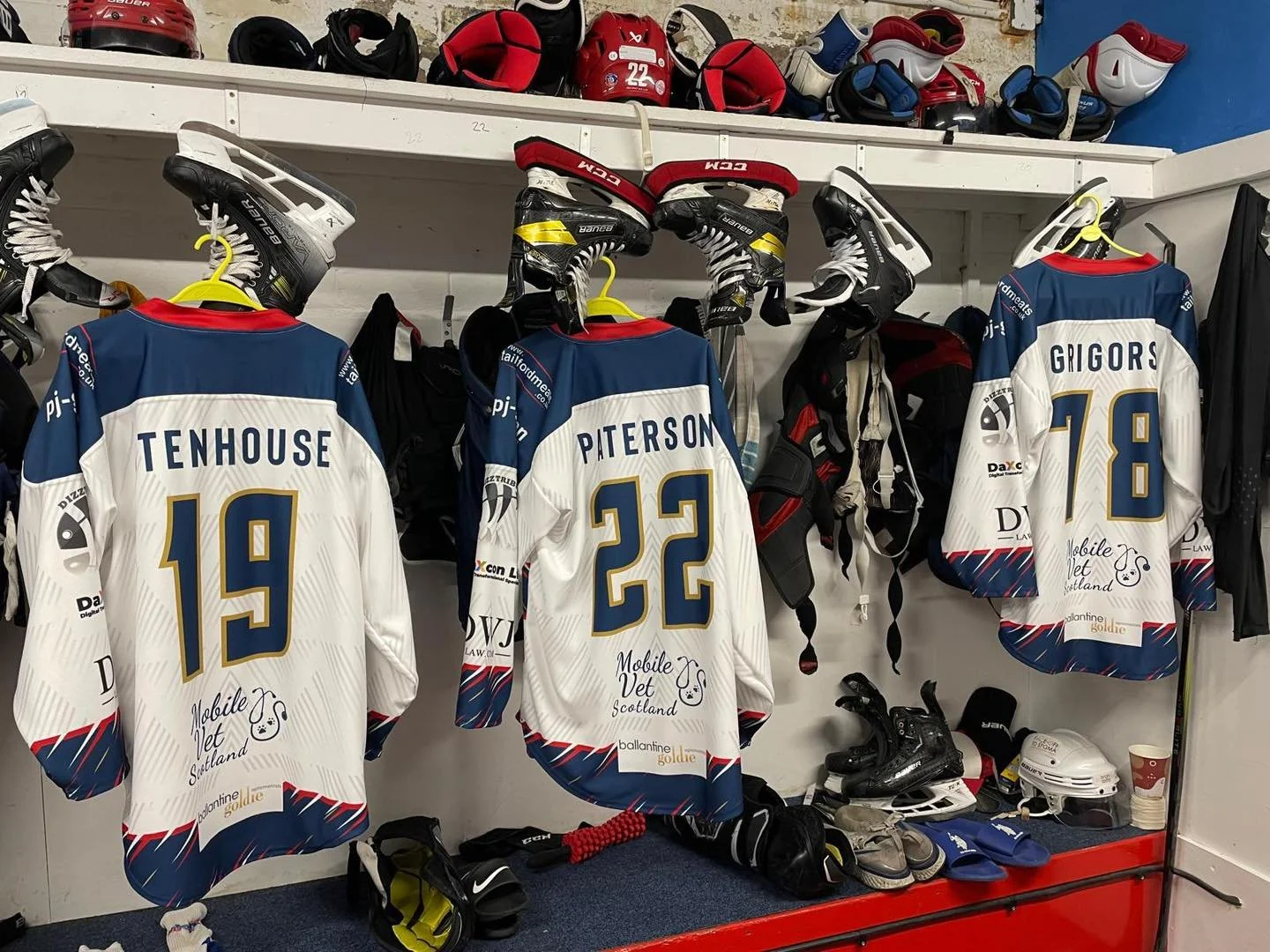 Ice hockey lockers painted white with three jerseys hanging on hooks, each with a number and name: 15 Tenhouse, 22 Paterson, 18 Grigos. Helmets, gloves, and skates are placed on shelves and the floor.