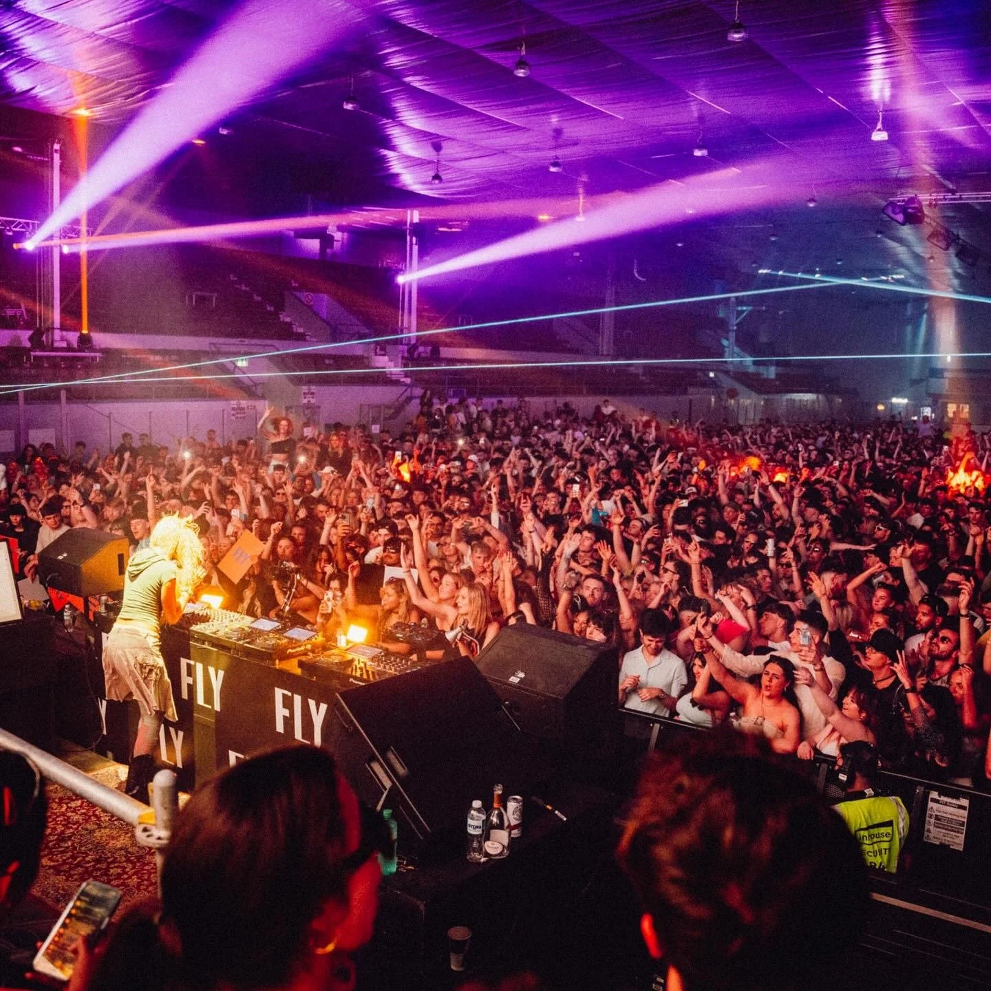 A crowded indoor concert with a DJ on stage, colorful lights and laser beams, and a large audience dancing and enjoying the music.