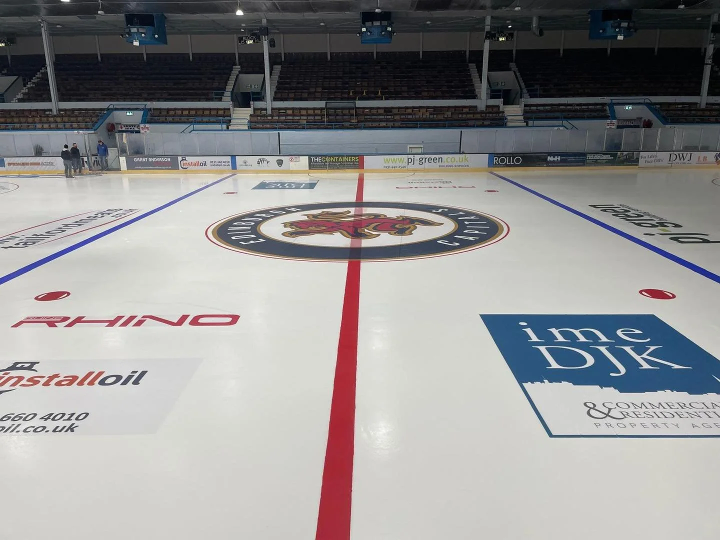 Empty indoor ice hockey rink with the team's logo at center, surrounded by advertising boards and seating areas.