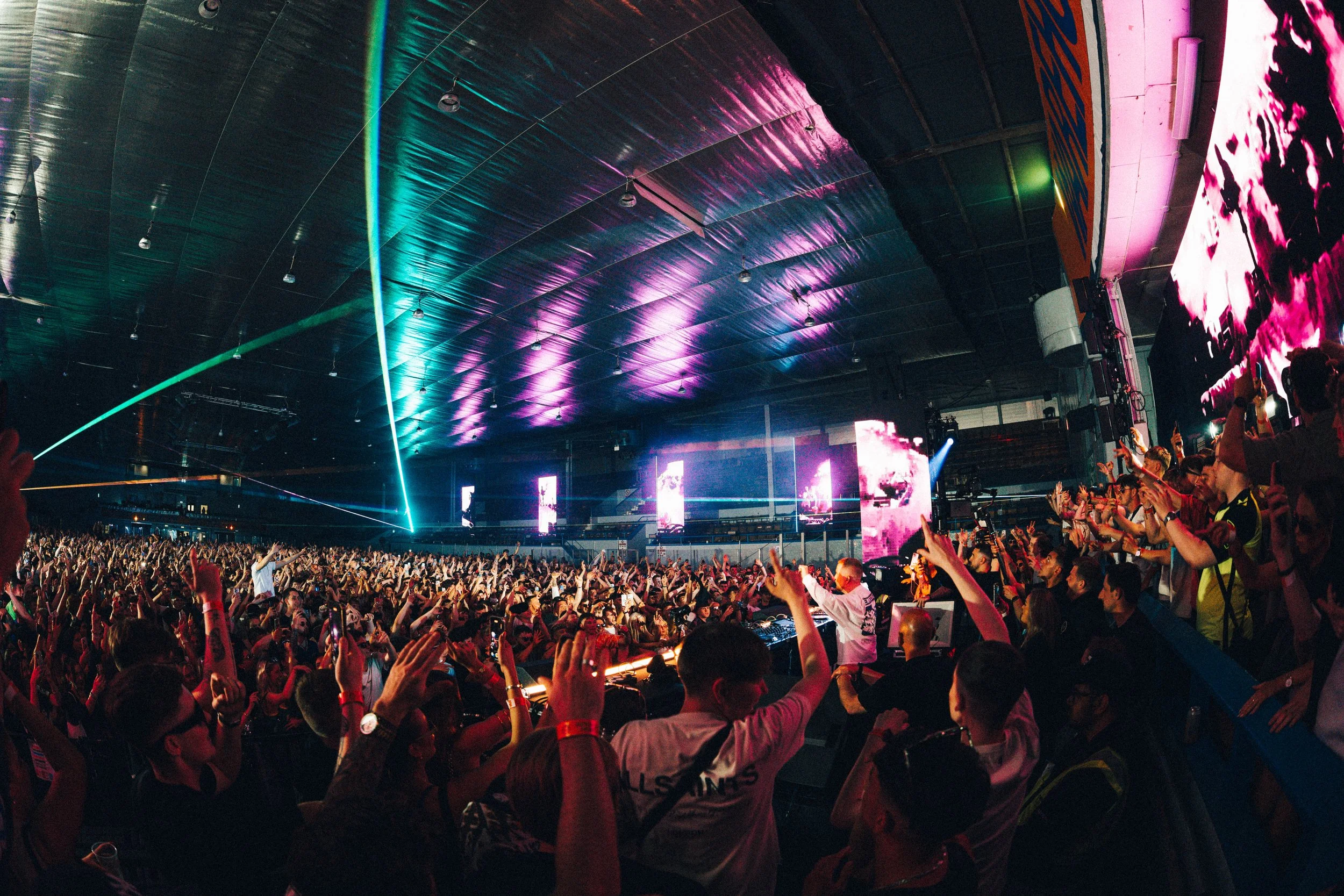 Large crowd at a concert or music event inside a spacious venue with colorful lights and laser beams.