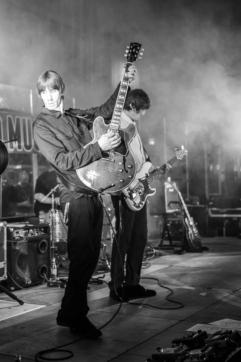 Black and white photo of two musicians performing on stage, one playing an electric guitar and the other playing an acoustic guitar, with audio equipment and microphone stands visible.