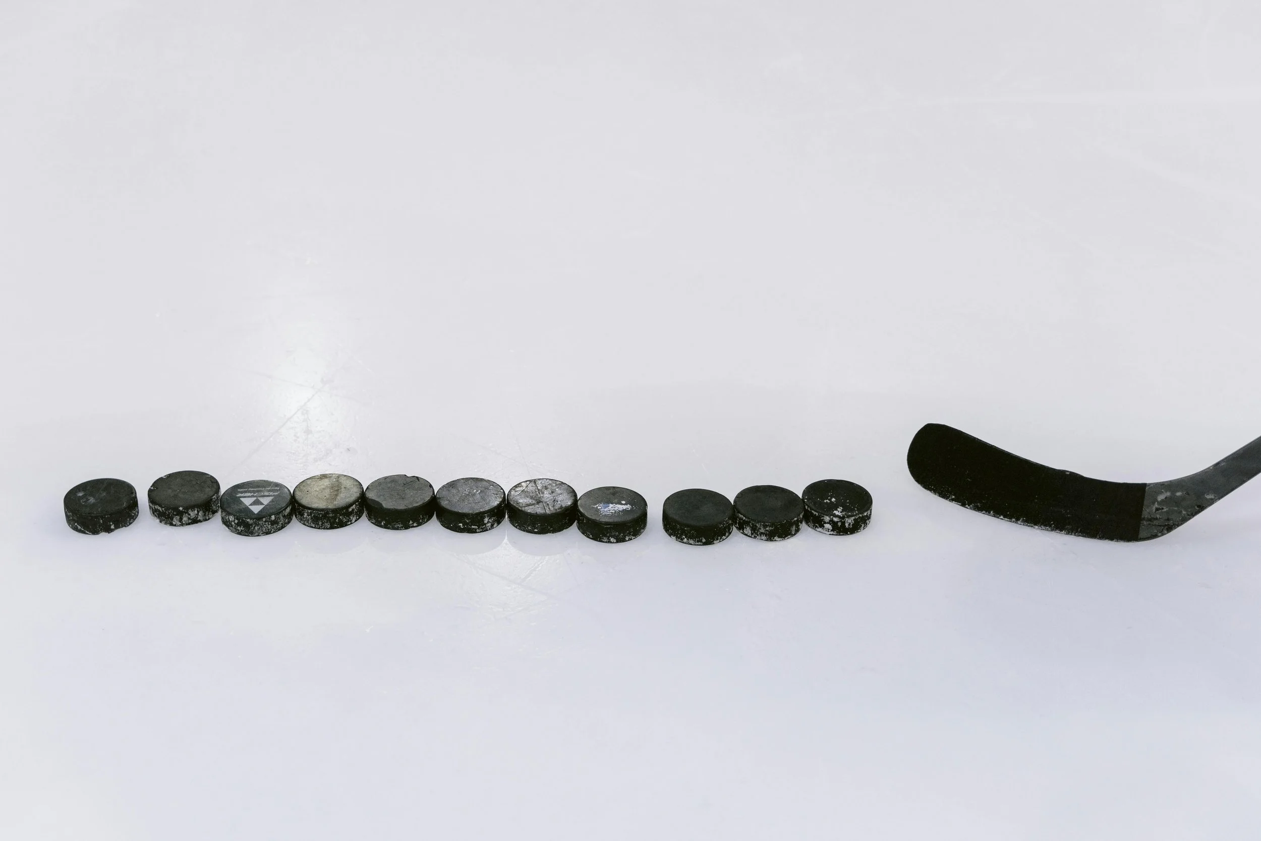 Line of black hockey pucks on a white ice surface with a hockey stick on the right.
