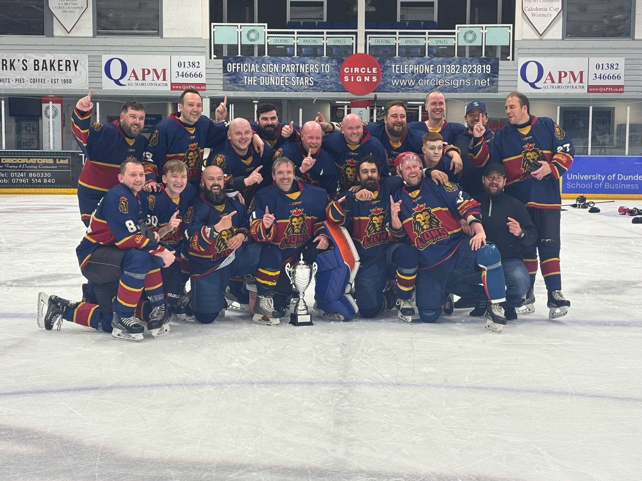 A group of ice hockey players in blue and maroon jerseys celebrating on an ice rink, holding a trophy and posing for a team photo.