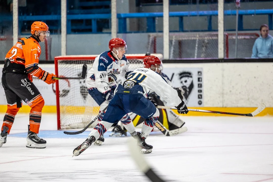 Ice hockey players in action during a game, with players in orange, white, and blue jerseys near the goal.