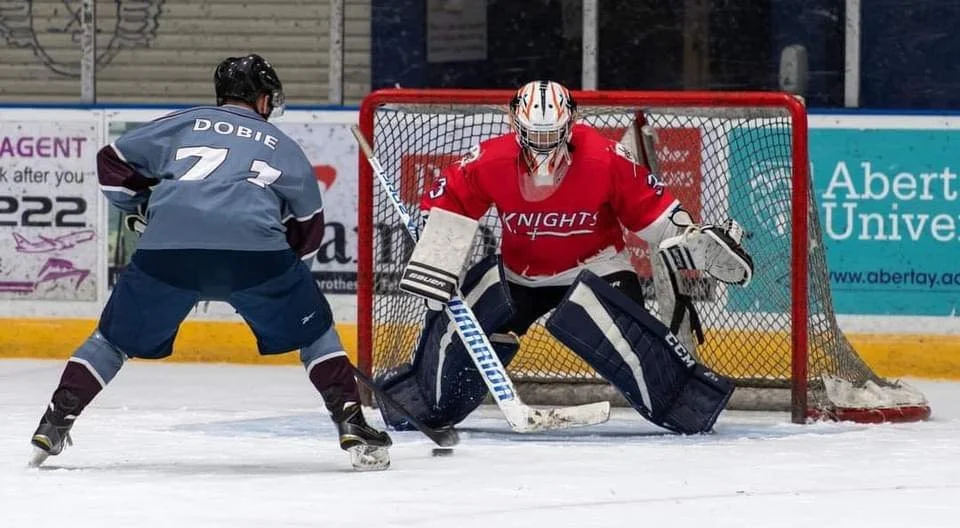 An ice hockey player wearing a grey jersey with the number 7 and the name Dobie faces a goalie in red and white gear, positioned in front of the net during a game.