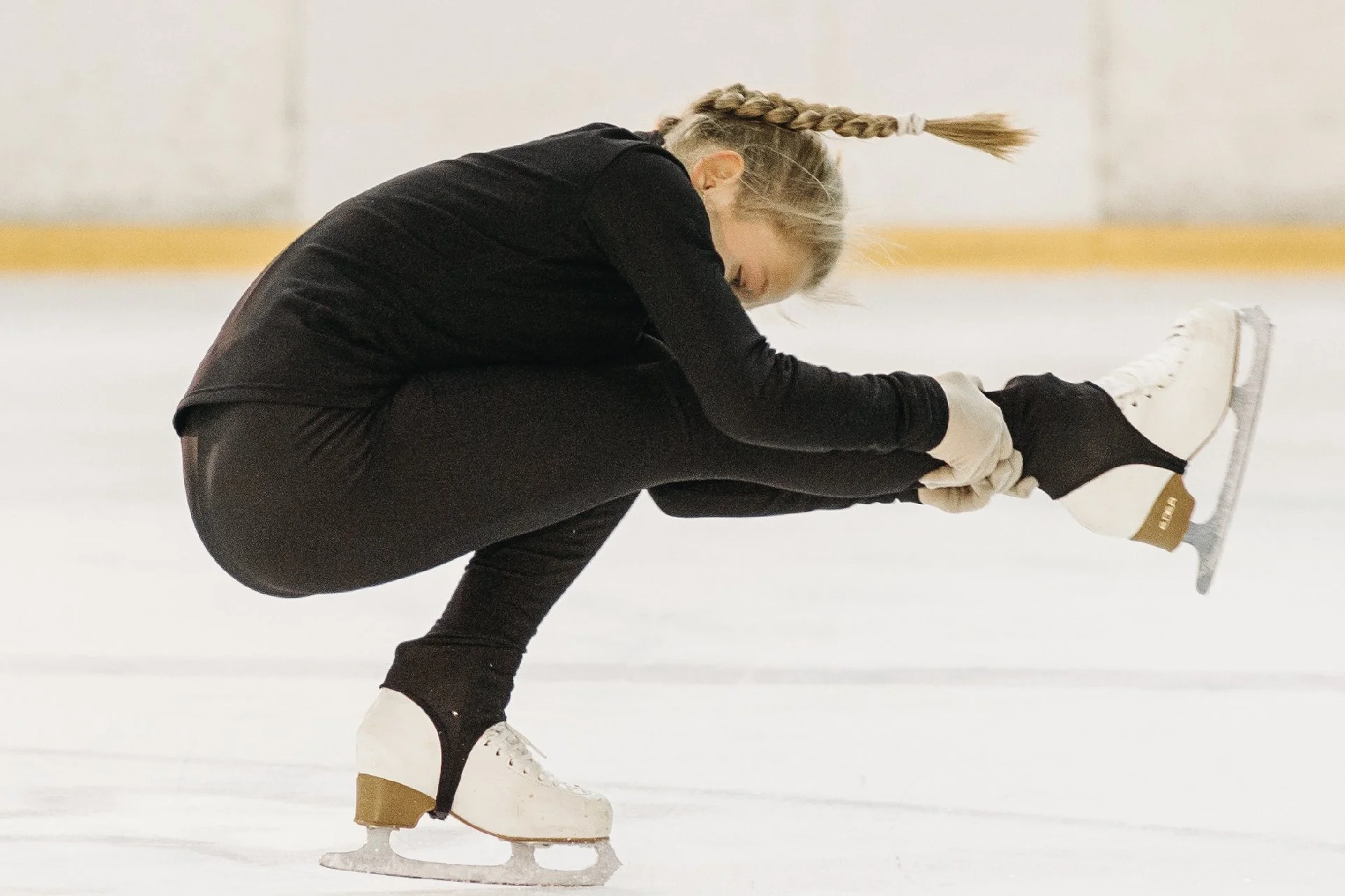 A female figure skater in black outfit, wearing white ice skates and gloves, is performing a move on the ice rink. She is bent forward, holding her right ankle with her right hand, and her left leg is extended backward. Her blonde hair is in a braid.
