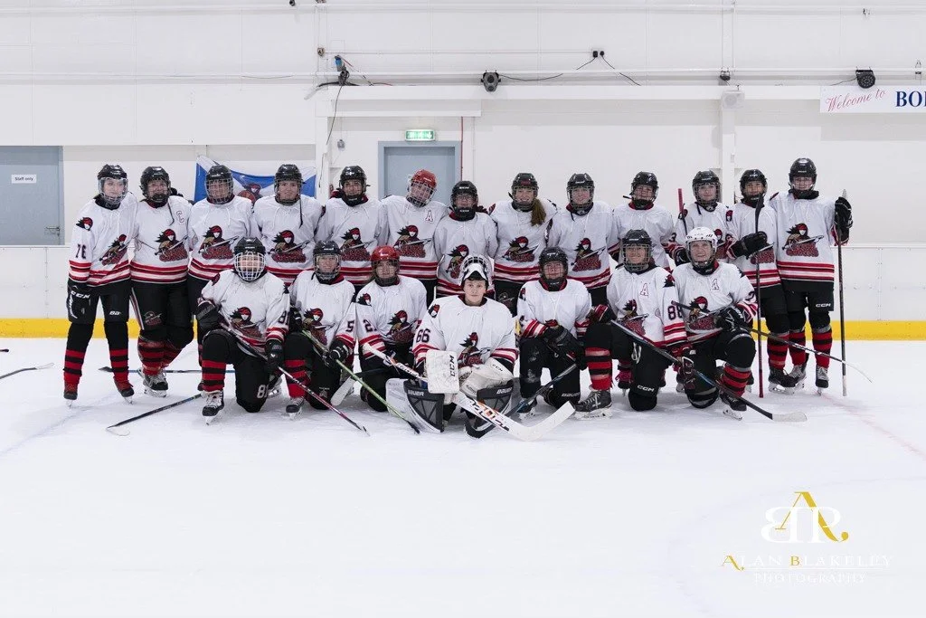 A group of young female ice hockey players wearing white jerseys with red and black accents, posing together on the ice rink.