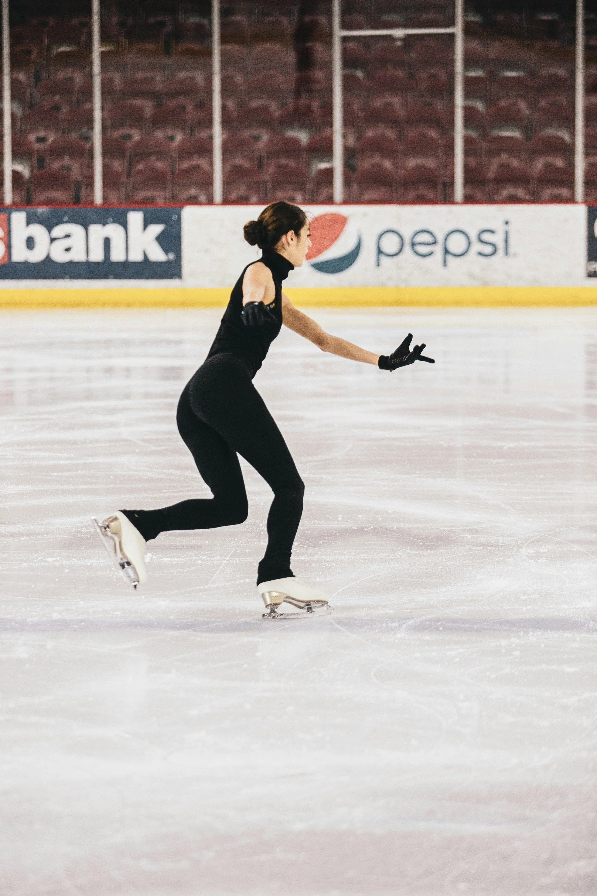 A female figure skater in black attire gliding on ice rink, with advertising banners in the background.