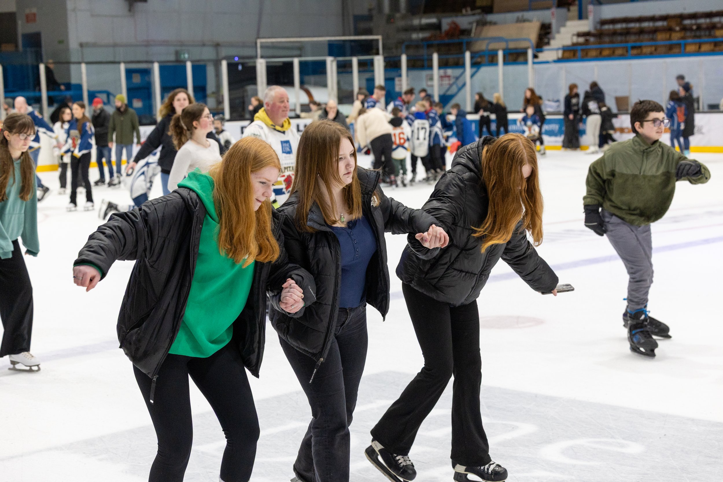 People ice skating at an indoor ice rink, holding hands and supporting each other