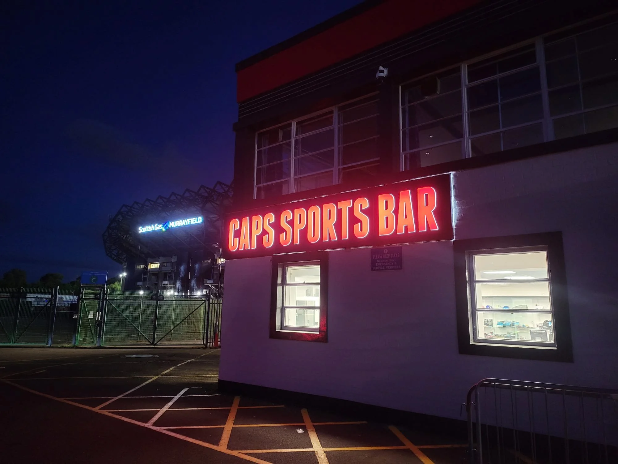 Night view of the exterior of a building with a large illuminated sign that reads 'CAPS SPORTS BAR'. Behind, there is a stadium with a sign that reads 'Scottish G…. MURAYFIELD' and a viewing area, indicating the location is near a sports stadium.