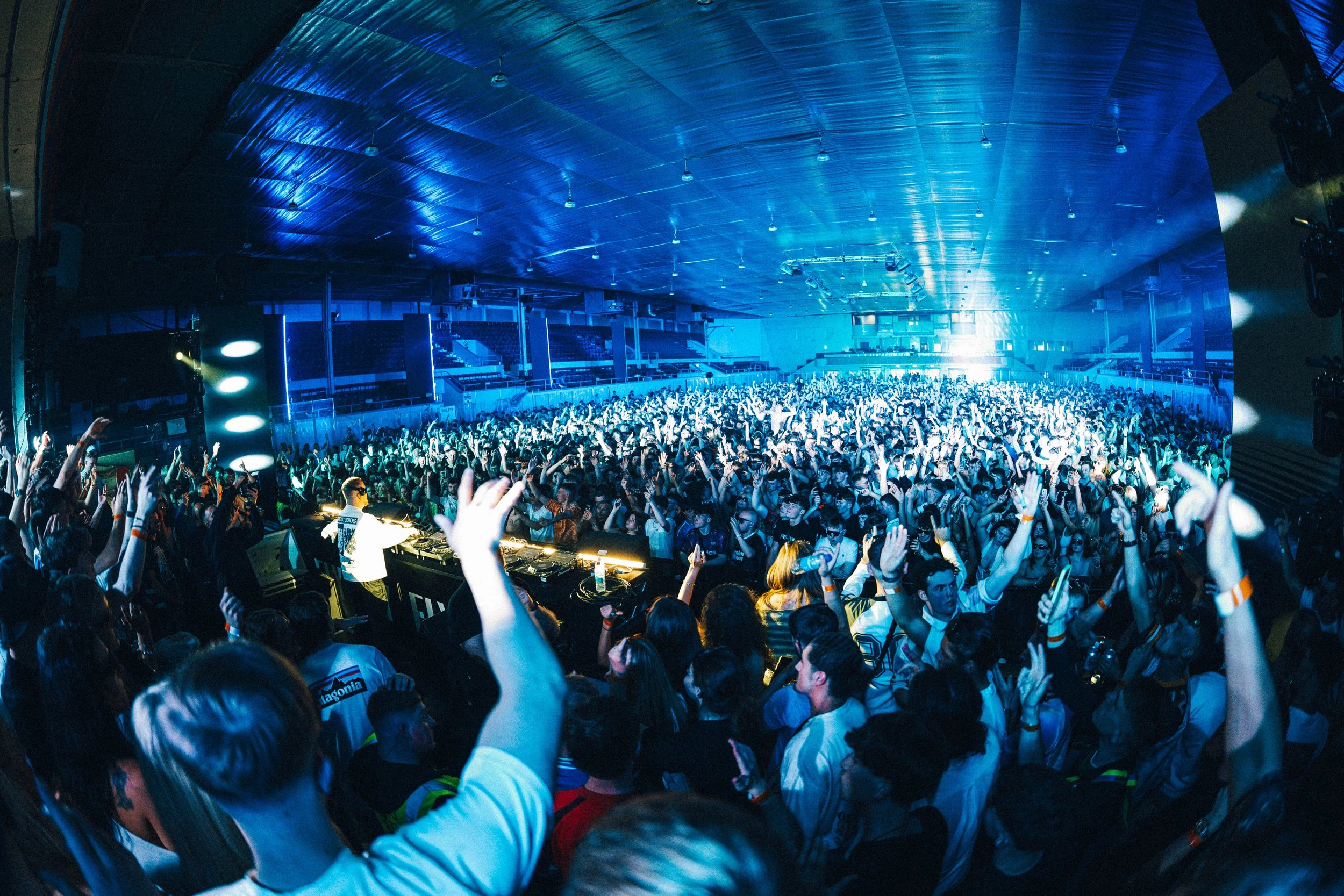 Crowd of people dancing and enjoying music at a concert or music festival inside an indoor arena with stage lights and DJ booth.